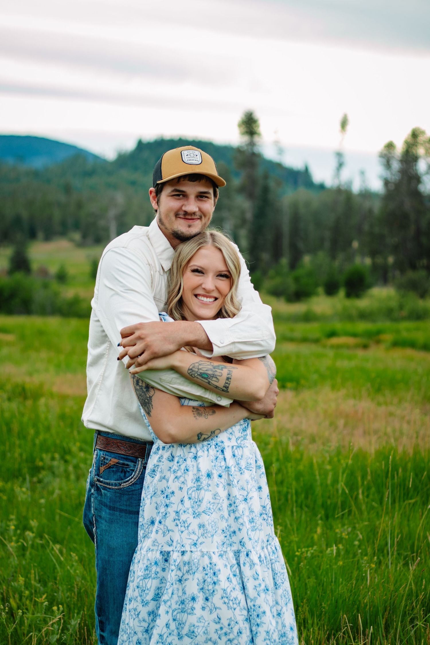 Happy couple hugging in a green meadow, woman smiling in floral dress, man in white shirt and cap.