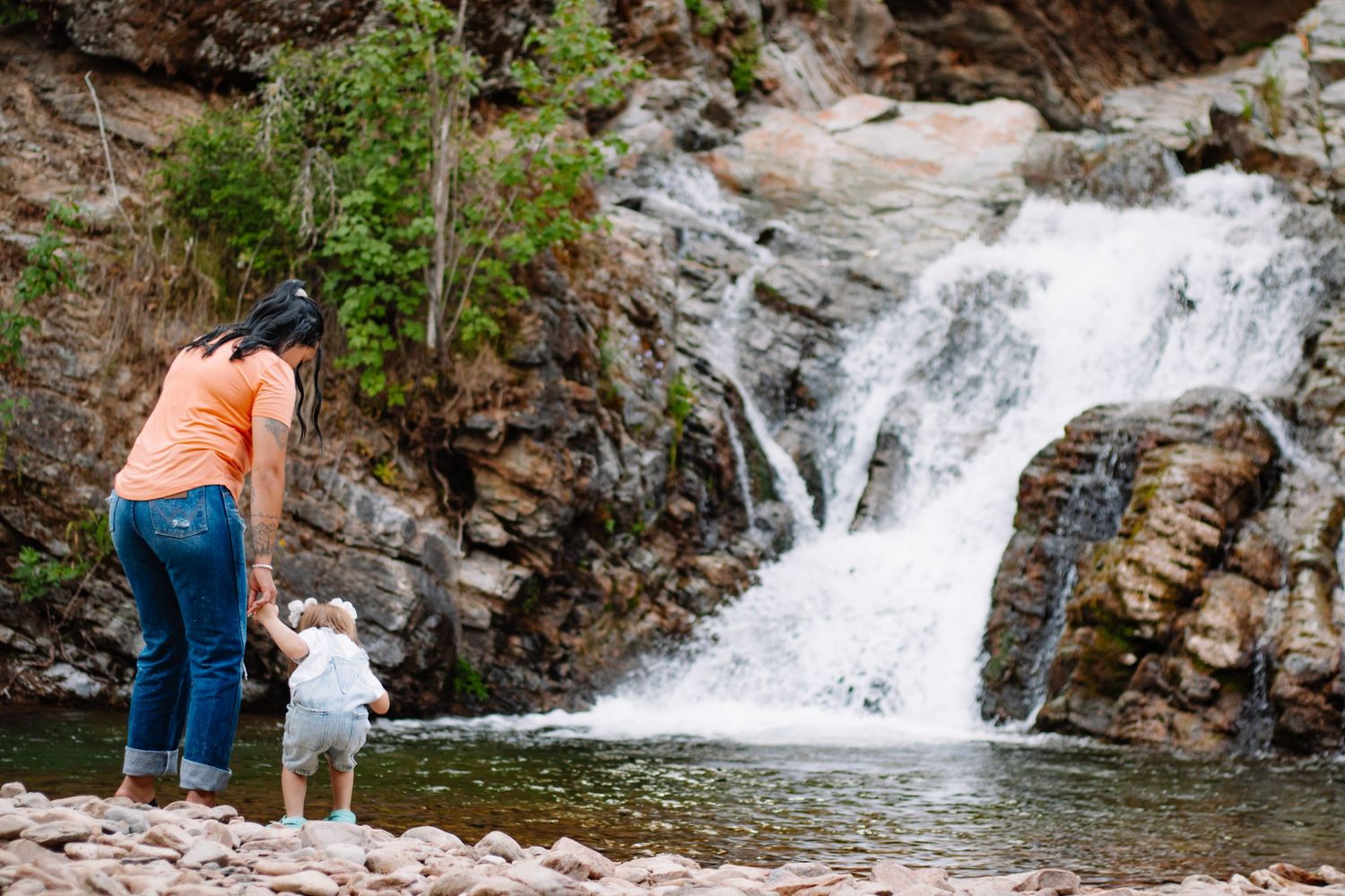 Mother and toddler exploring rocky shoreline beside a scenic cascading waterfall surrounded by lush greenery.