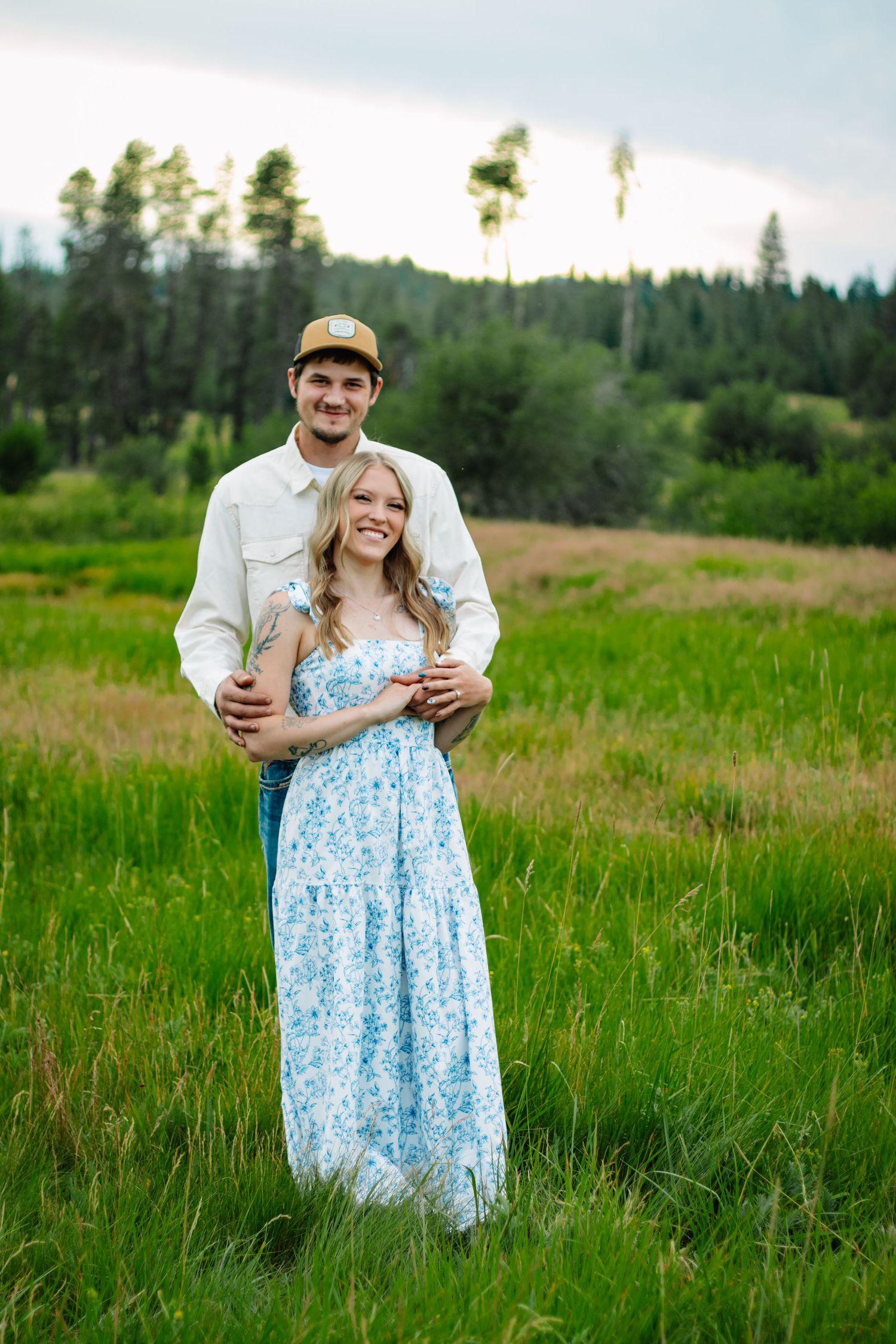 Couple embracing in a lush green meadow, man in white shirt and cap hugging woman in blue floral dress.
