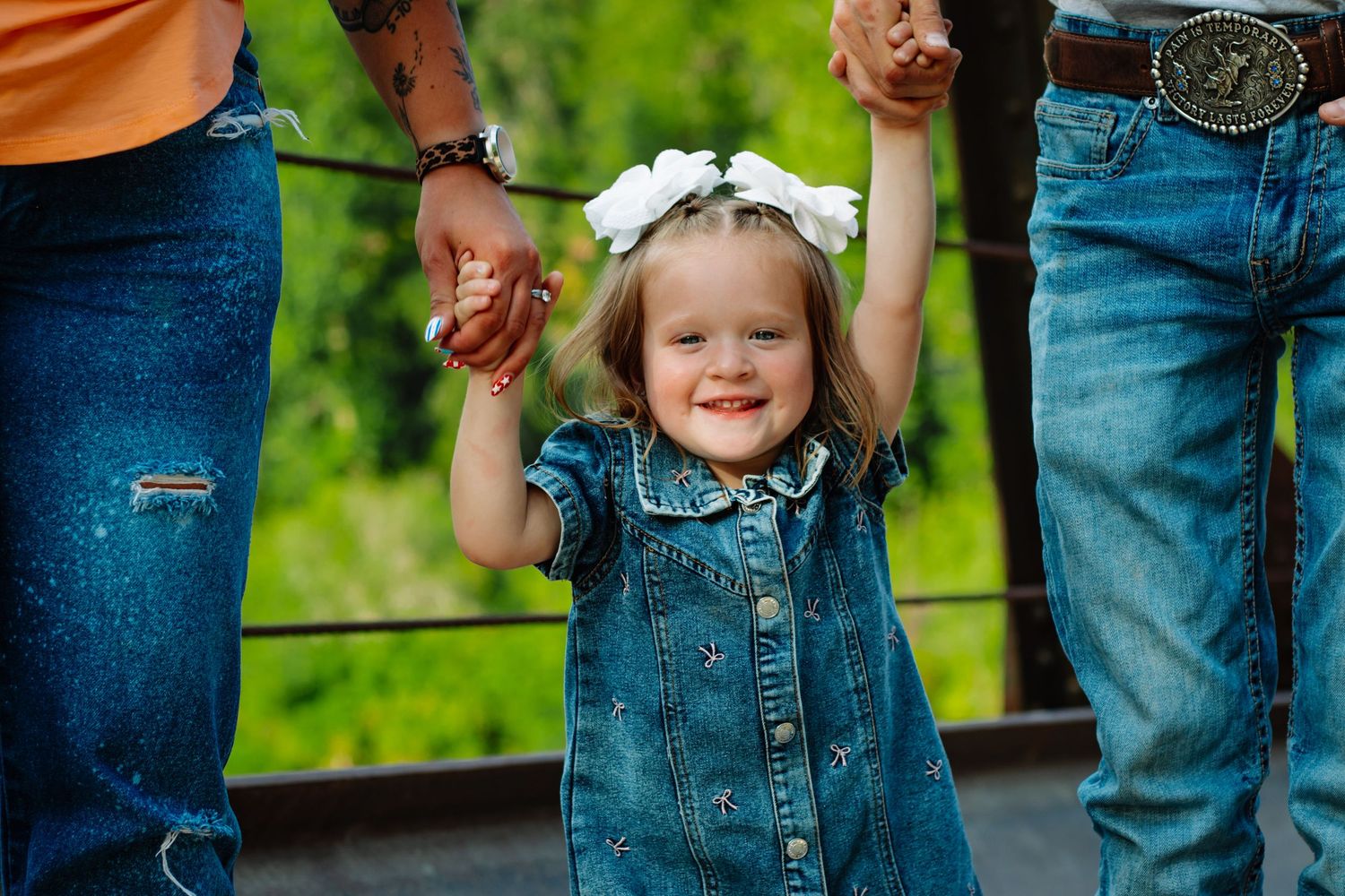 Happy toddler girl in denim dress holding parents hands on outdoor bridge.