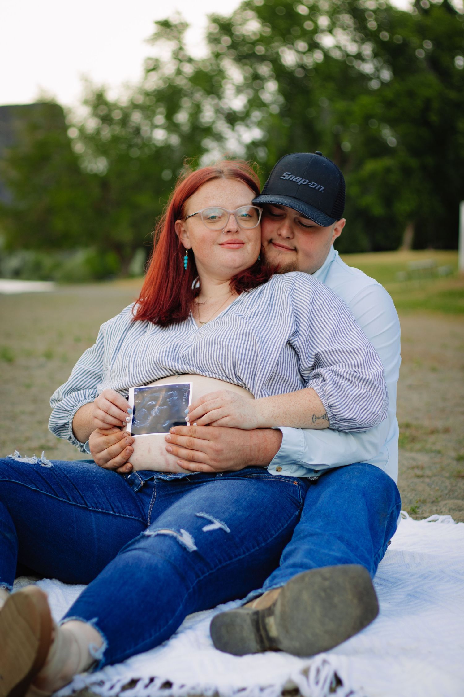 Couple sitting on blanket outdoors holding ultrasound photo, pregnancy announcement in park setting.