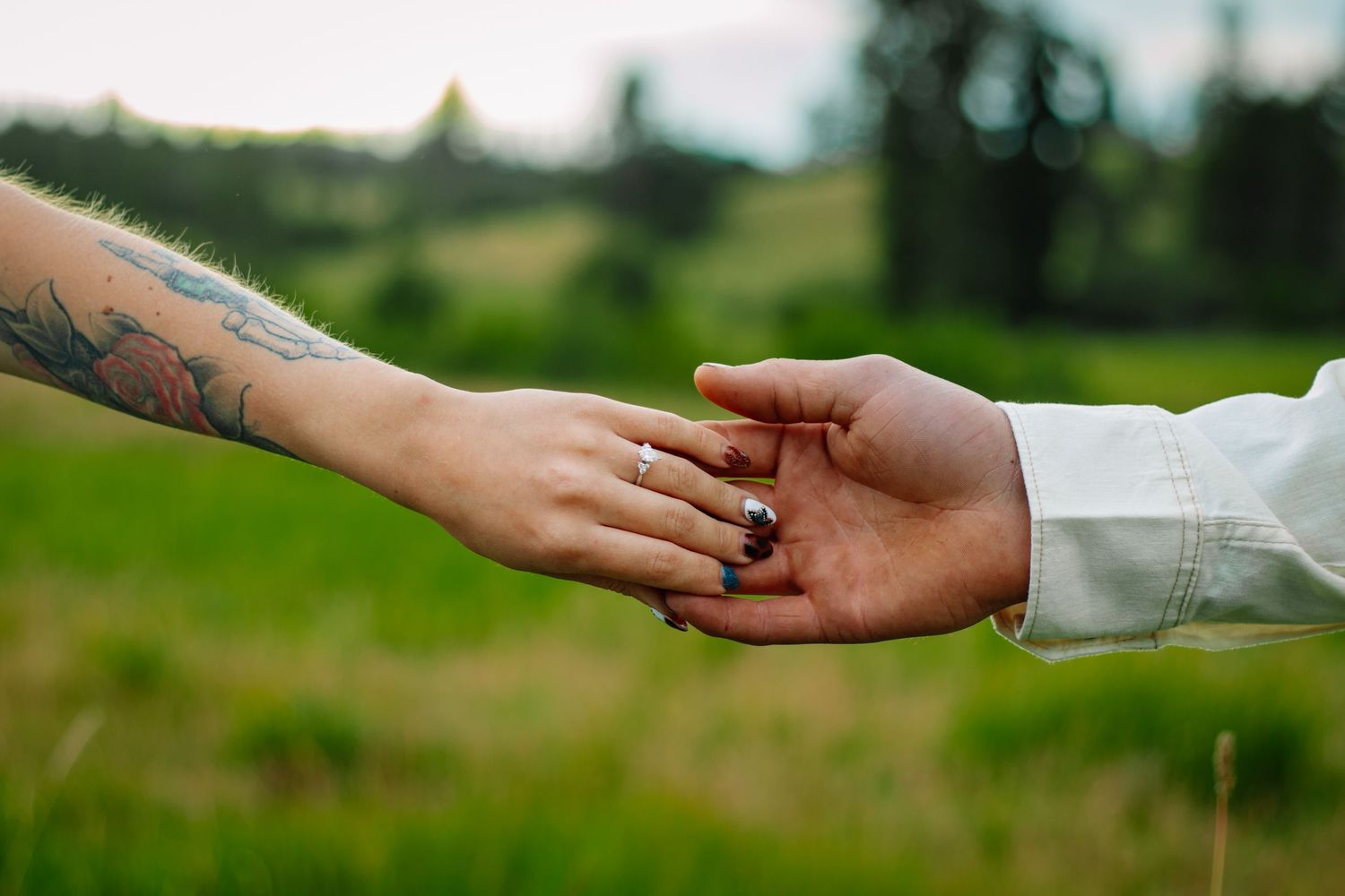 Close-up of couple holding hands in a green field, woman with rose tattoo wearing engagement ring.