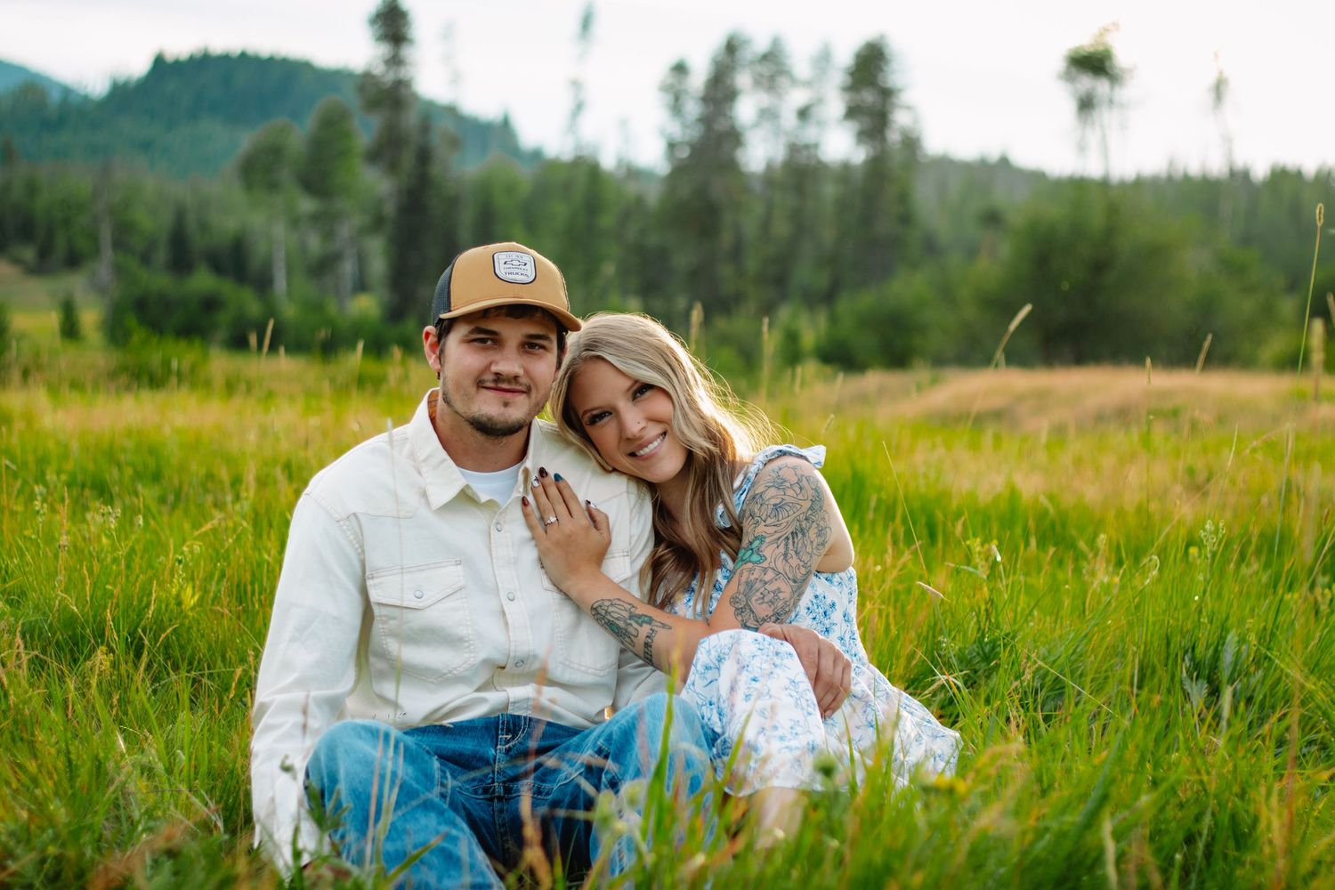 Couple sitting together in tall green meadow grass, woman leaning on mans shoulder smiling softly.