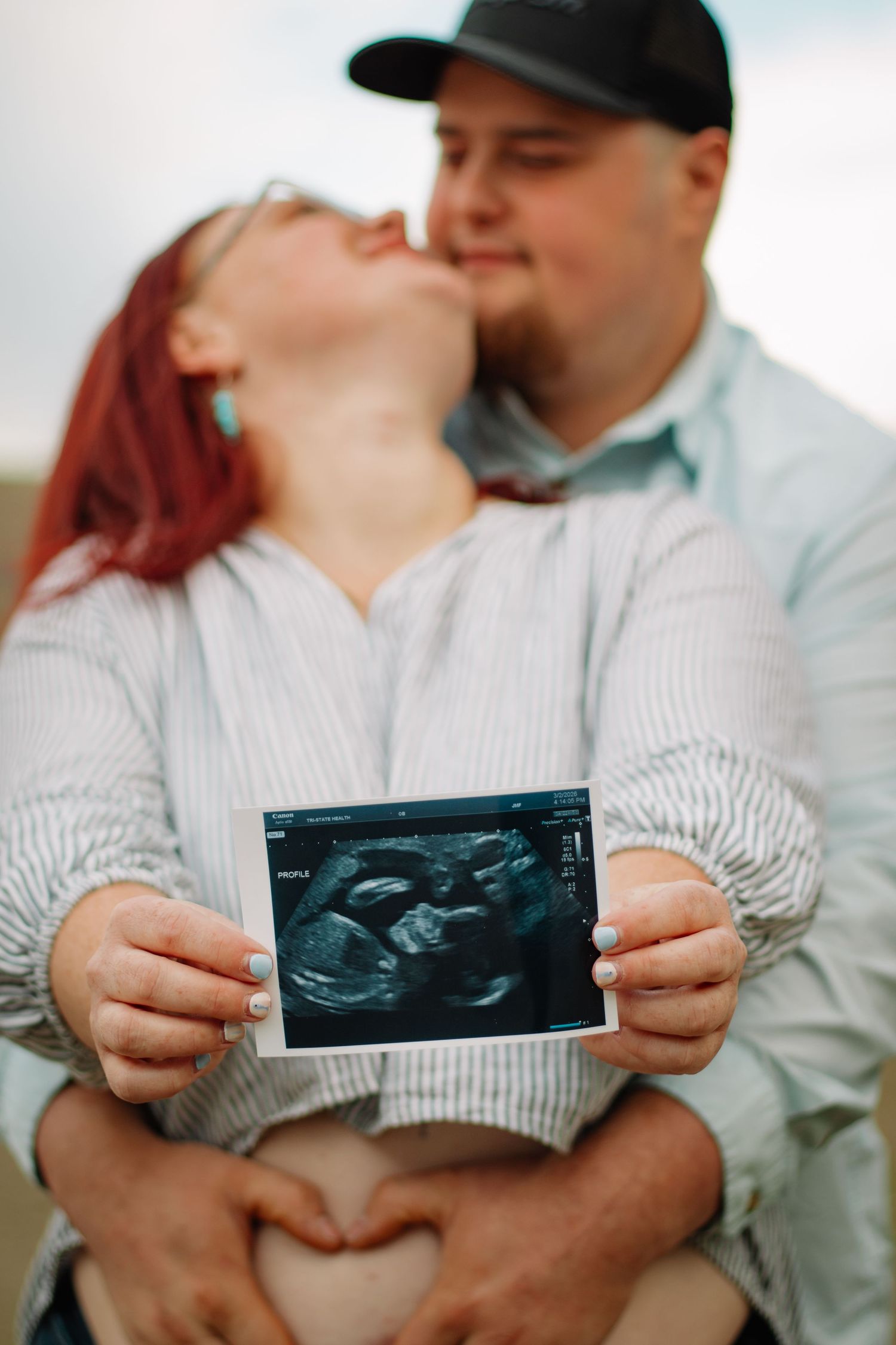 Couple kissing while holding ultrasound photo and making heart shape on baby bump.