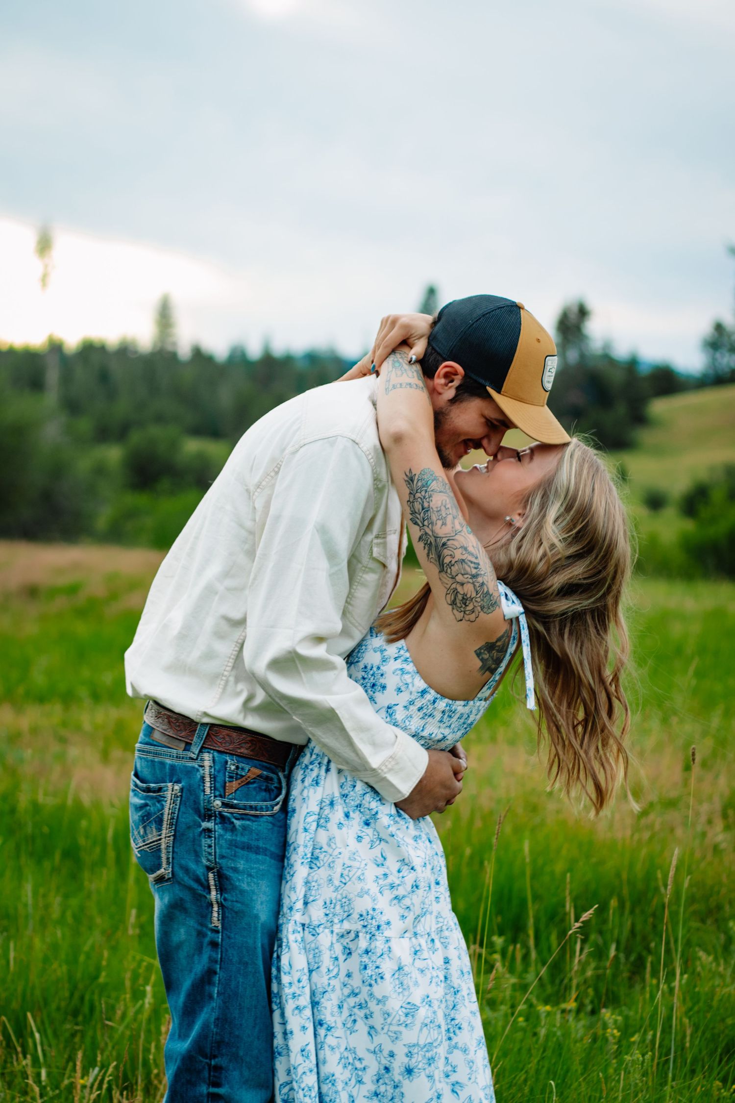 Couple sharing a romantic kiss in a green field, woman in floral dress with tattoos, man in cap.