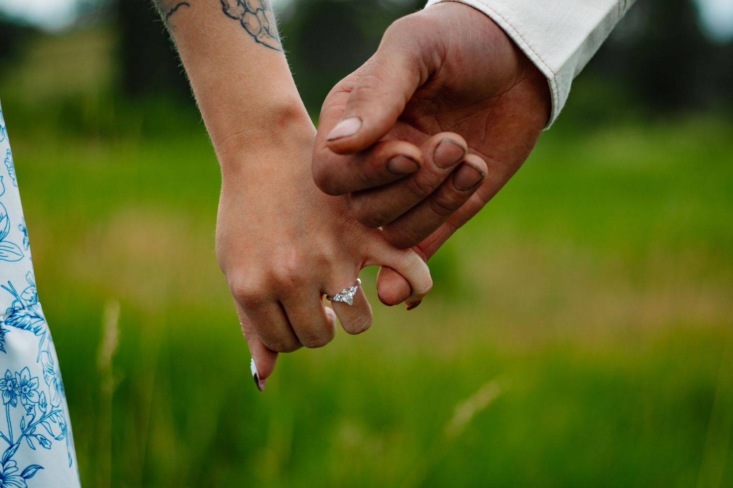 Close-up of couple holding hands showing engagement ring, woman with floral tattoo on wrist.