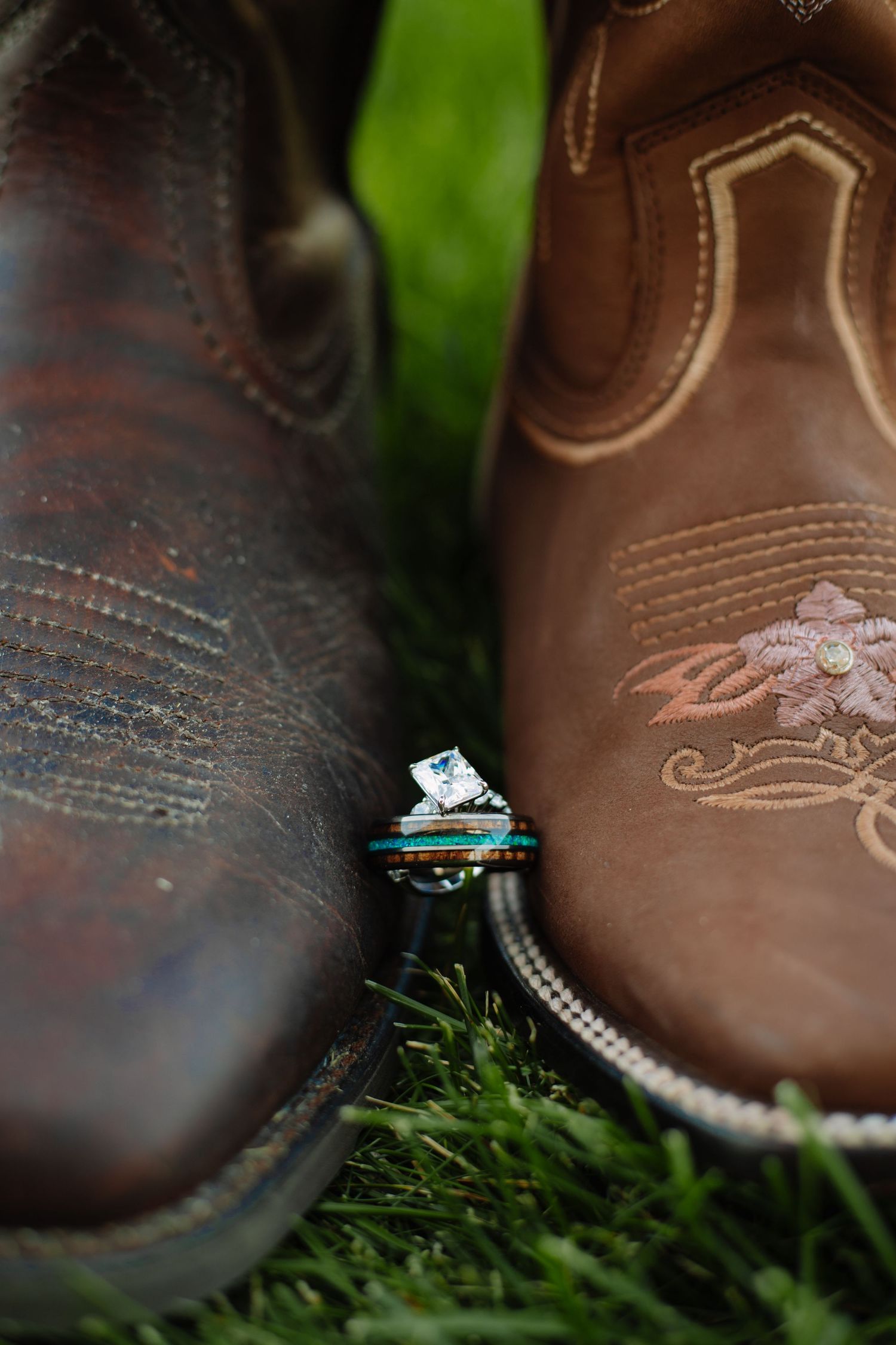 Wedding rings nestled between brown cowboy boots on green grass, symbolizing a rustic country wedding celebration.