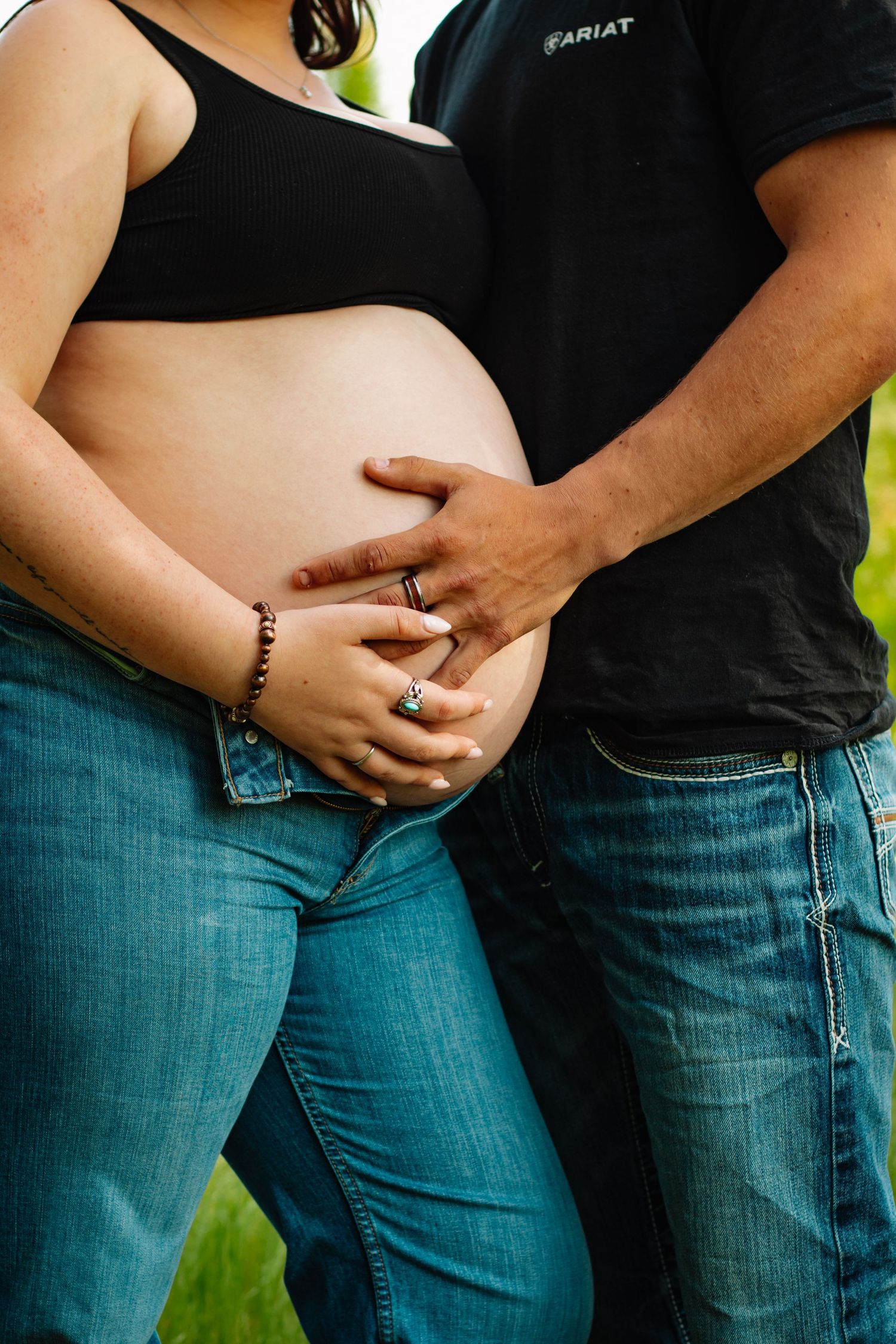 Couple holding pregnant belly together outdoors, wearing jeans and casual tops.