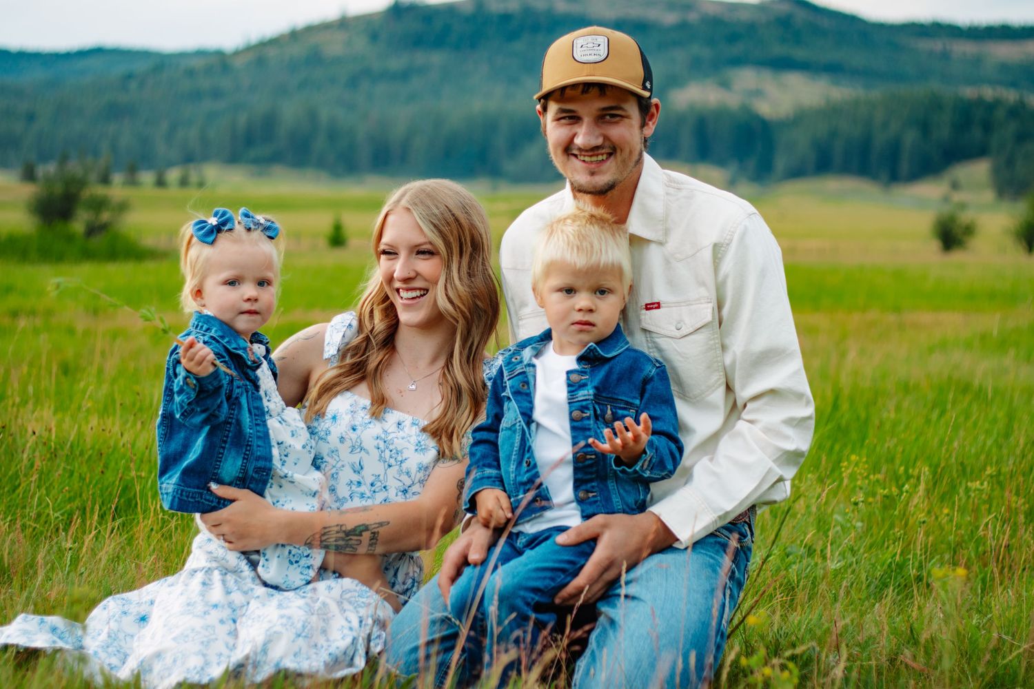 Family of four with toddlers in matching denim sitting together in scenic green meadow.
