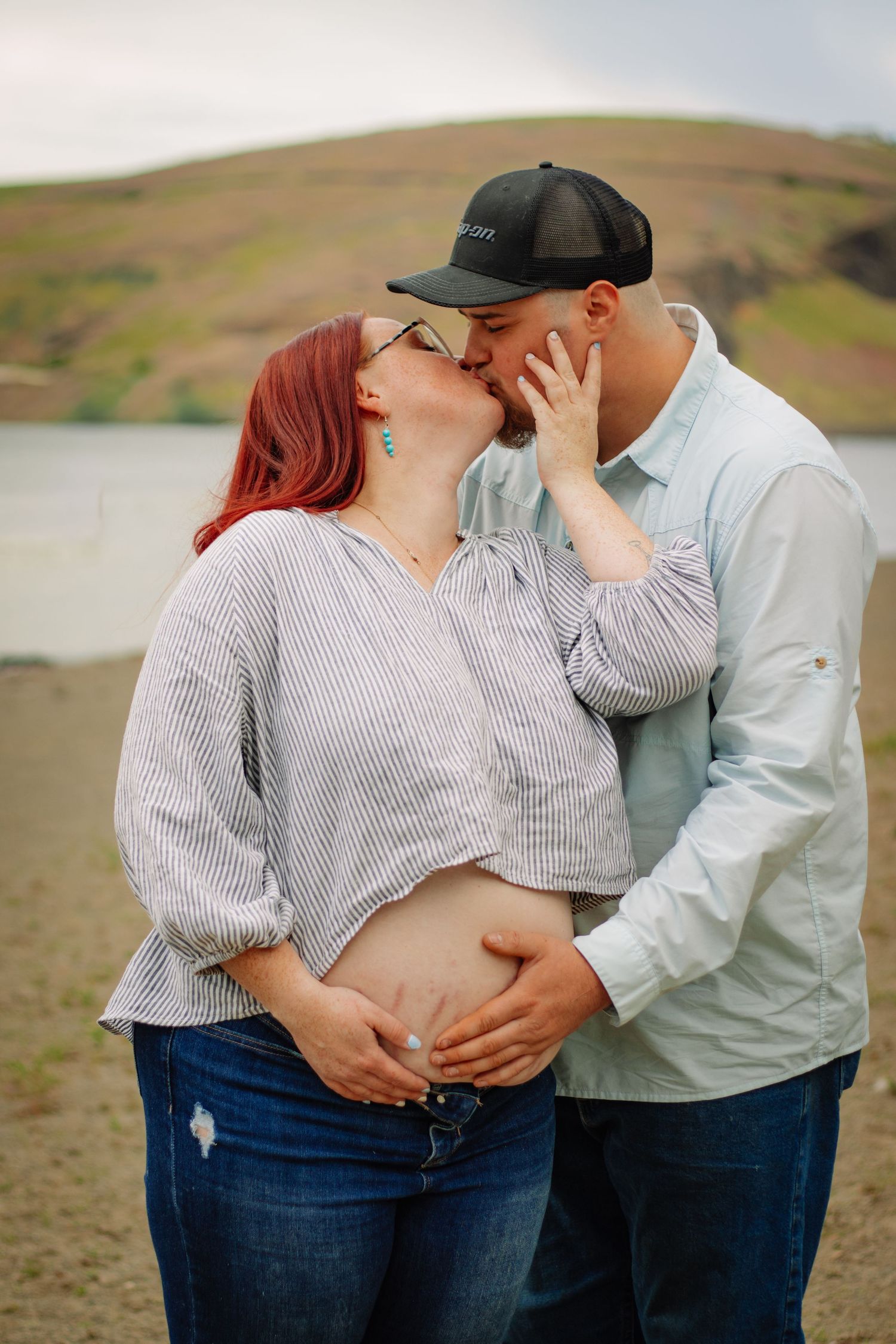 Pregnant redhead woman kissing partner near a lake, both cradling her baby bump.