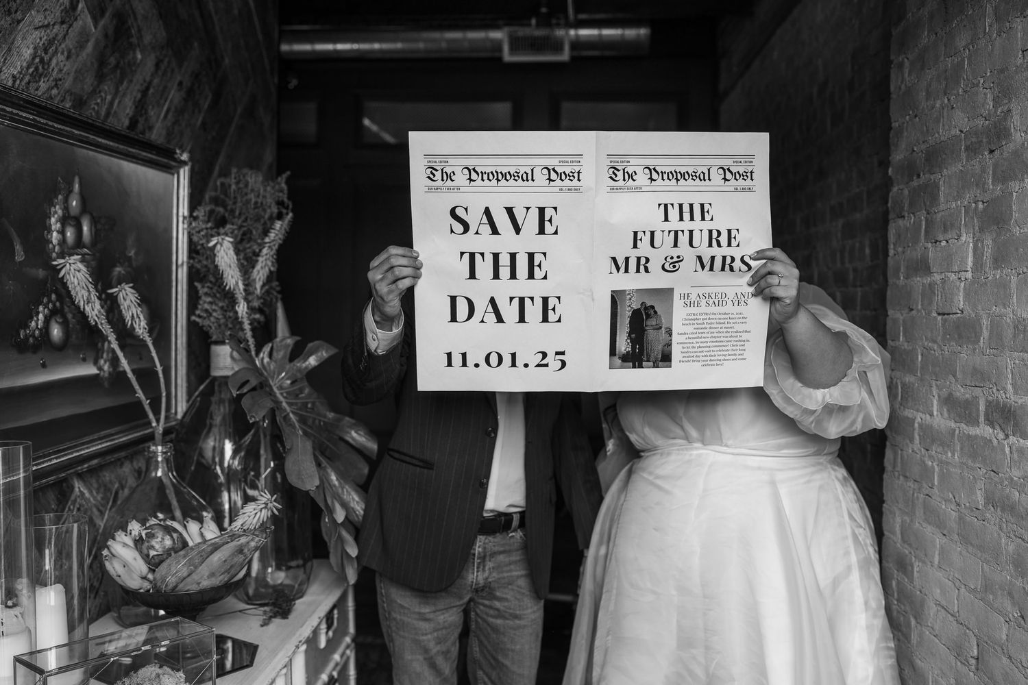 A couple holds creative save-the-date newspaper-style signs in black and white.