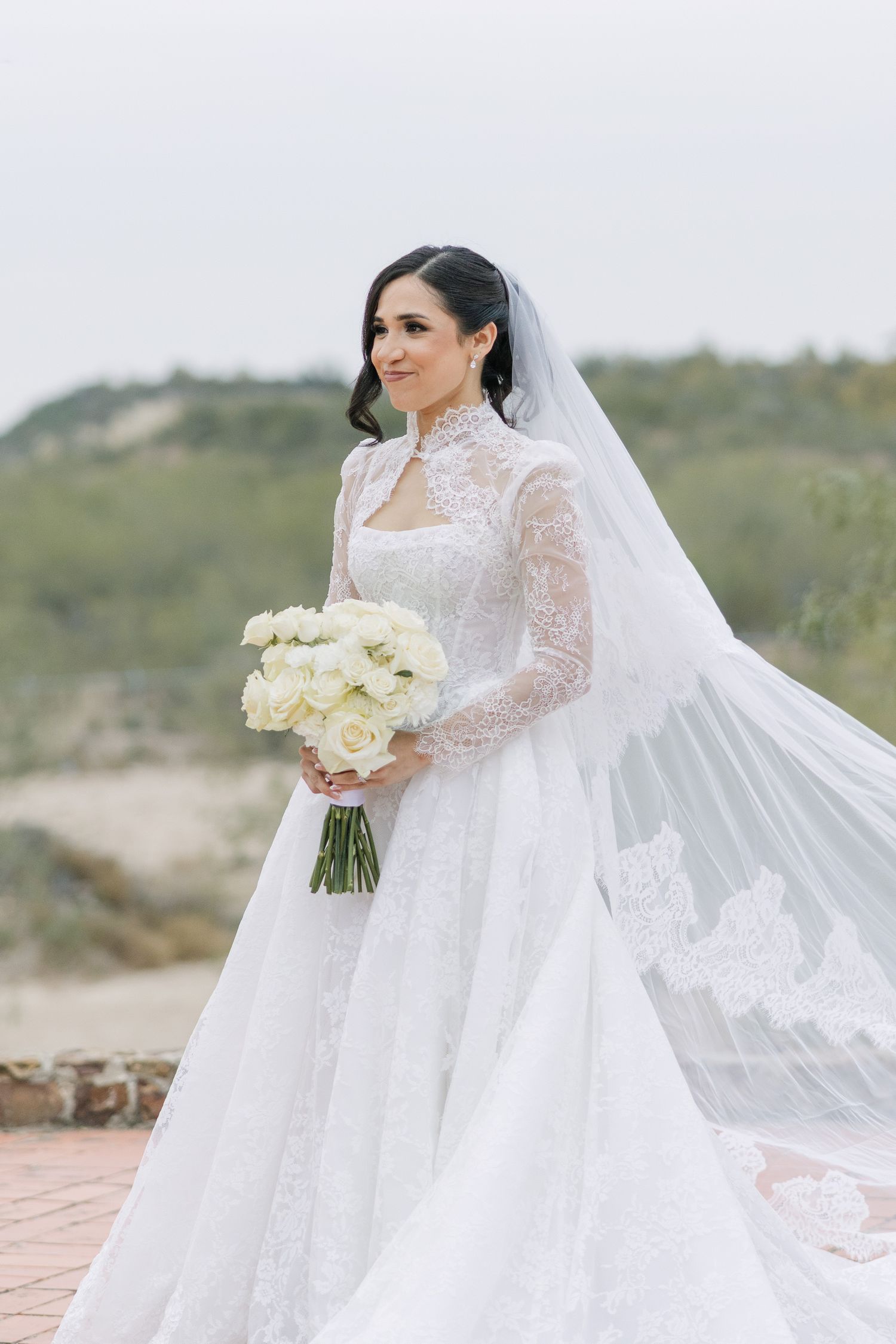 Bride in white lace long-sleeve wedding gown with veil holding white rose bouquet outdoors.