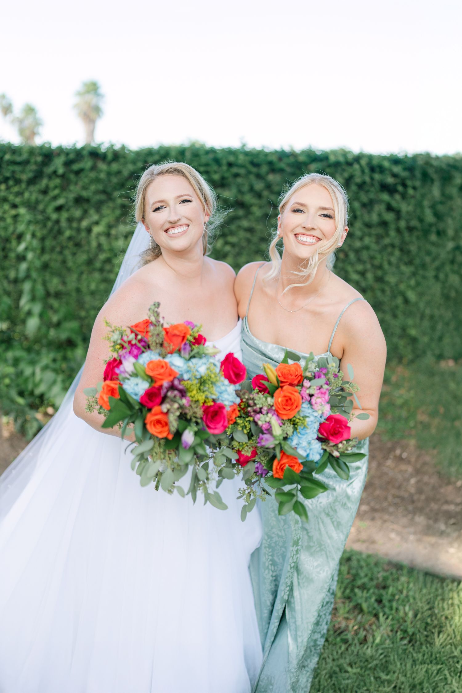Bride in white gown and bridesmaid in sage green dress holding vibrant floral bouquets in front of hedge wall.