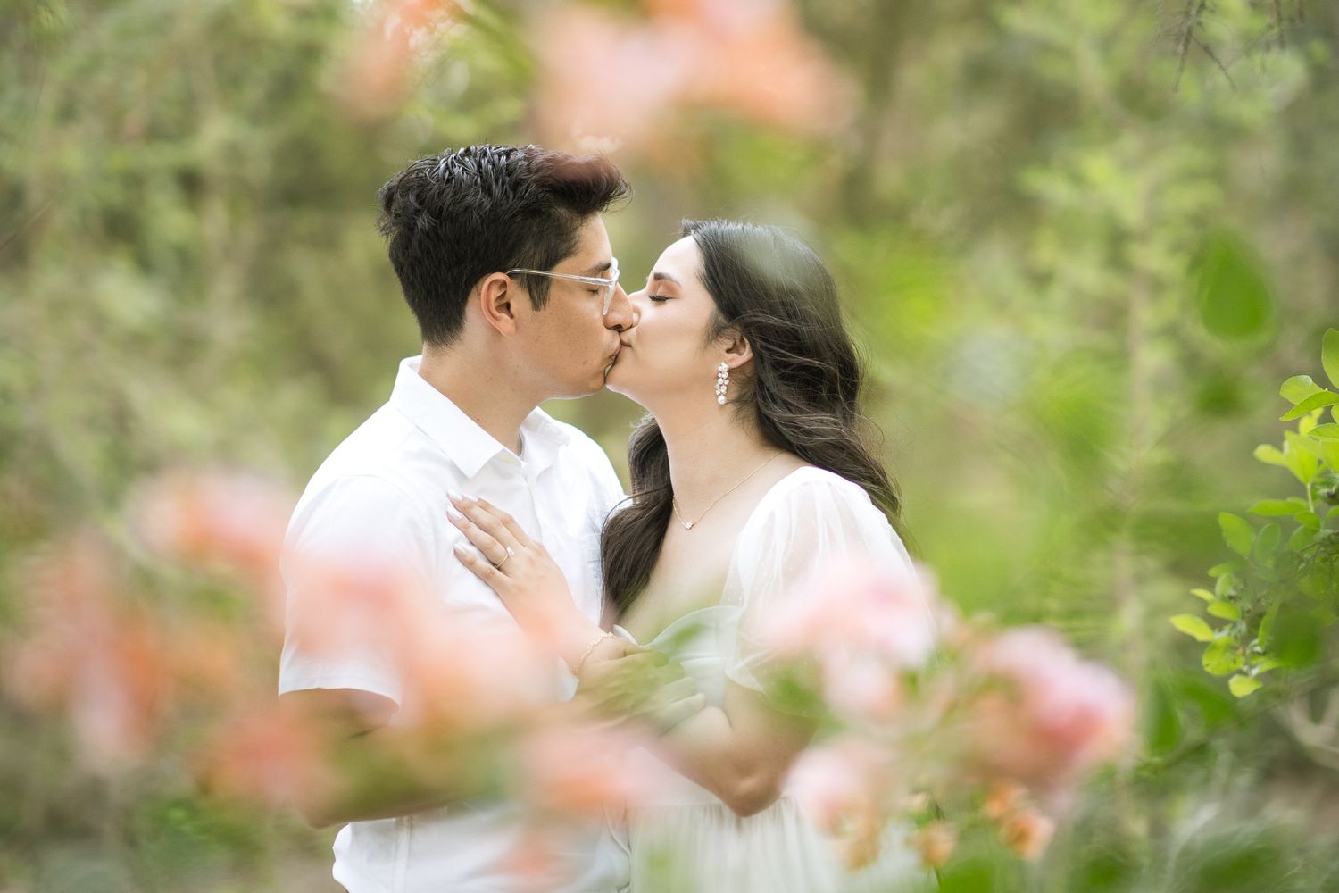 Two people share an intimate moment amid blooming pink flowers in a garden setting.