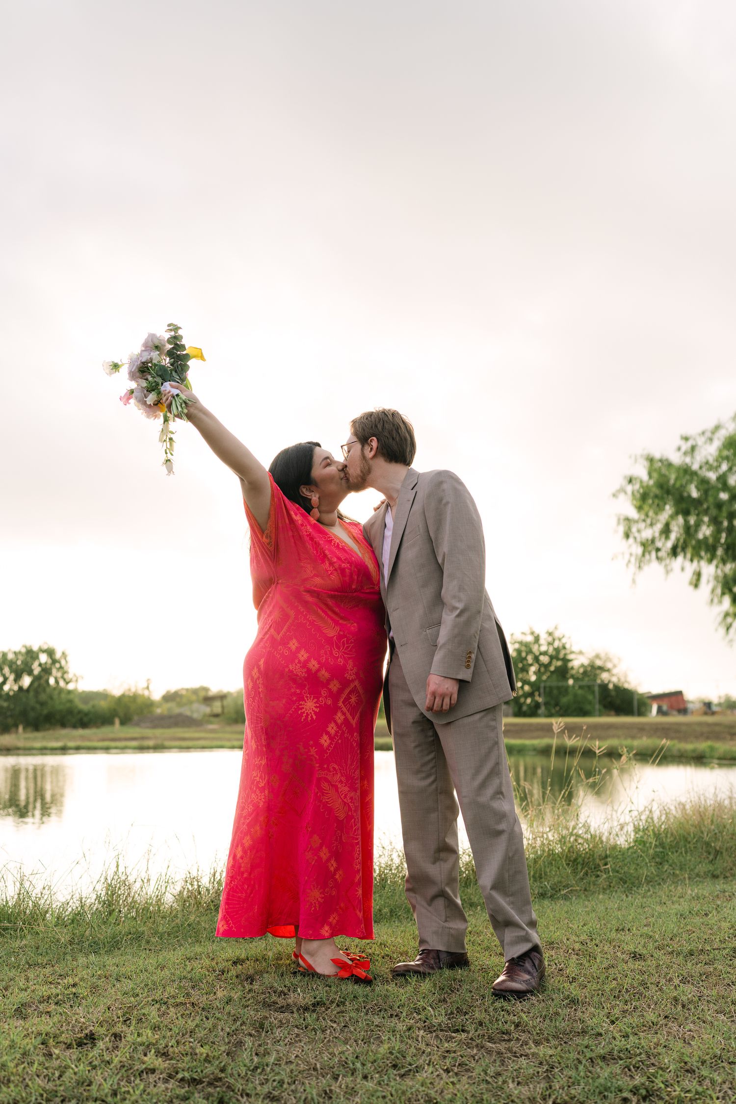 A couple shares a kiss by a lake while holding a bouquet against a sunset sky.