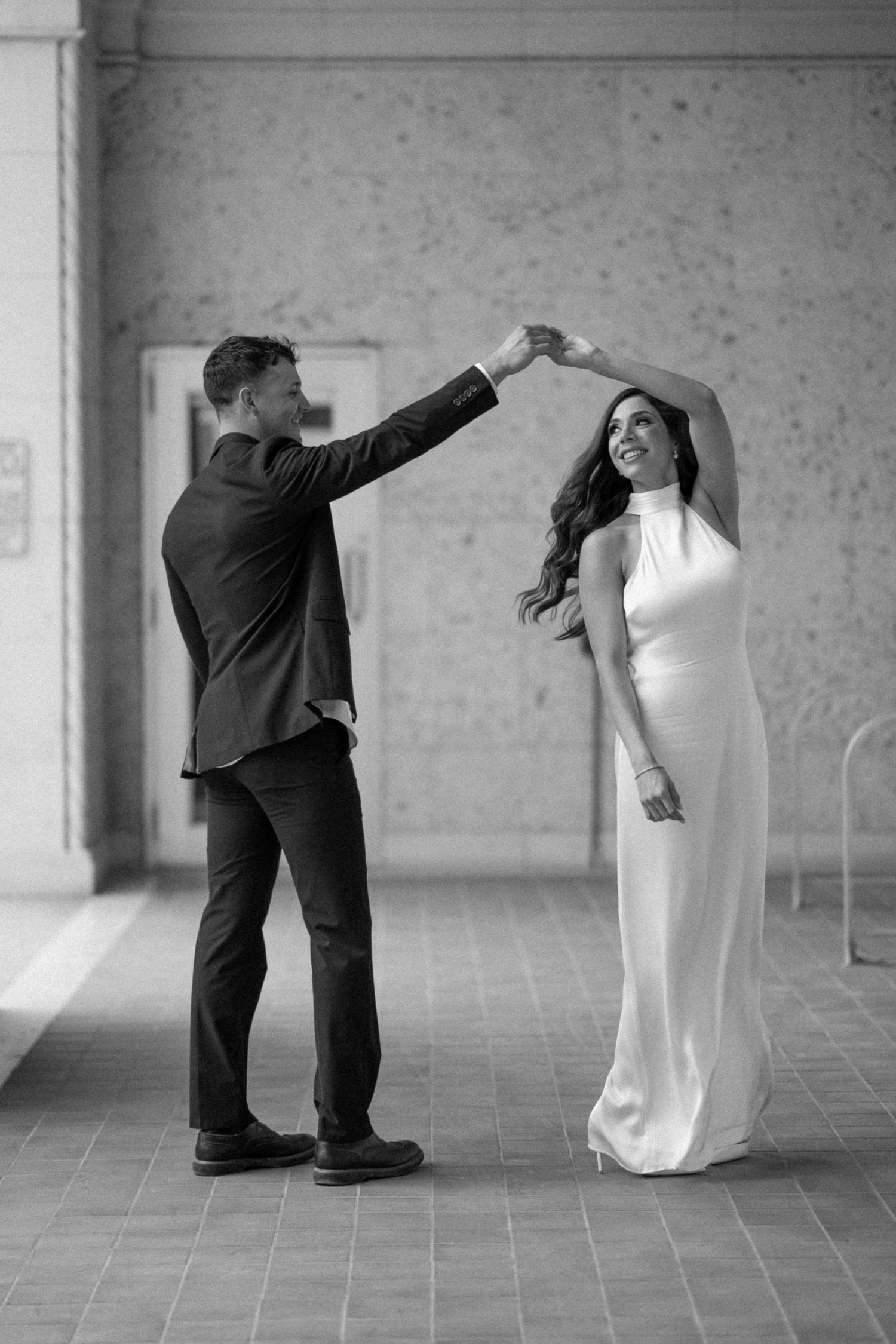 A couple in formal attire shares a playful dance move in a classic corridor.