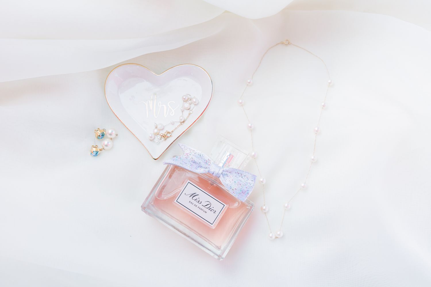 A delicate perfume bottle and jewelry arranged on soft white fabric with natural lighting.