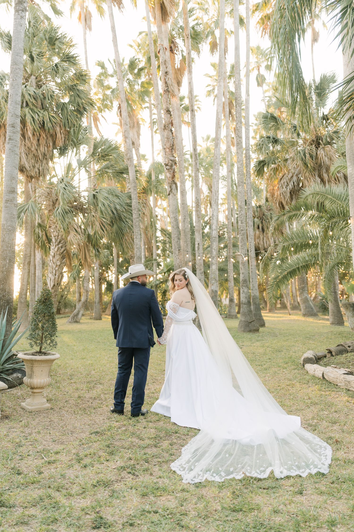 Bride and groom hold hands among tall palm trees, her lace cathedral veil trailing on lush green grass at golden hour.