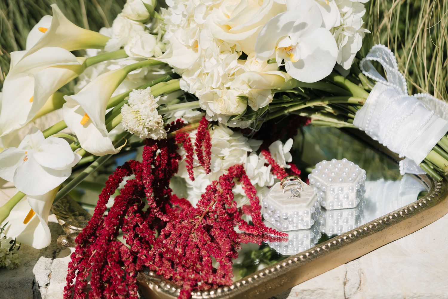 Elegant wedding bouquet with white calla lilies, orchids, and red amaranthus on a mirrored tray with jewelry boxes.