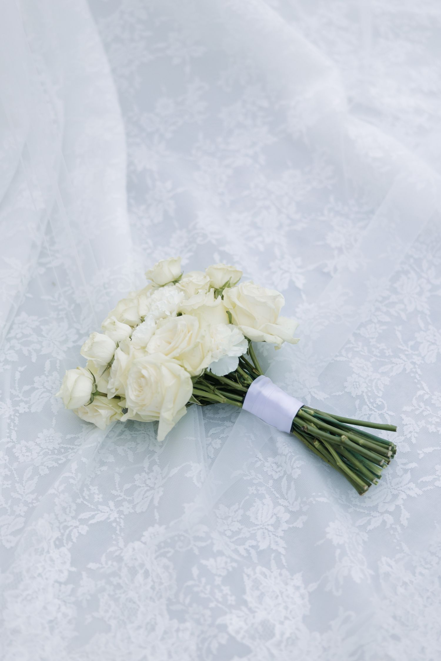 White rose bridal bouquet tied with ribbon resting on lace wedding dress fabric.