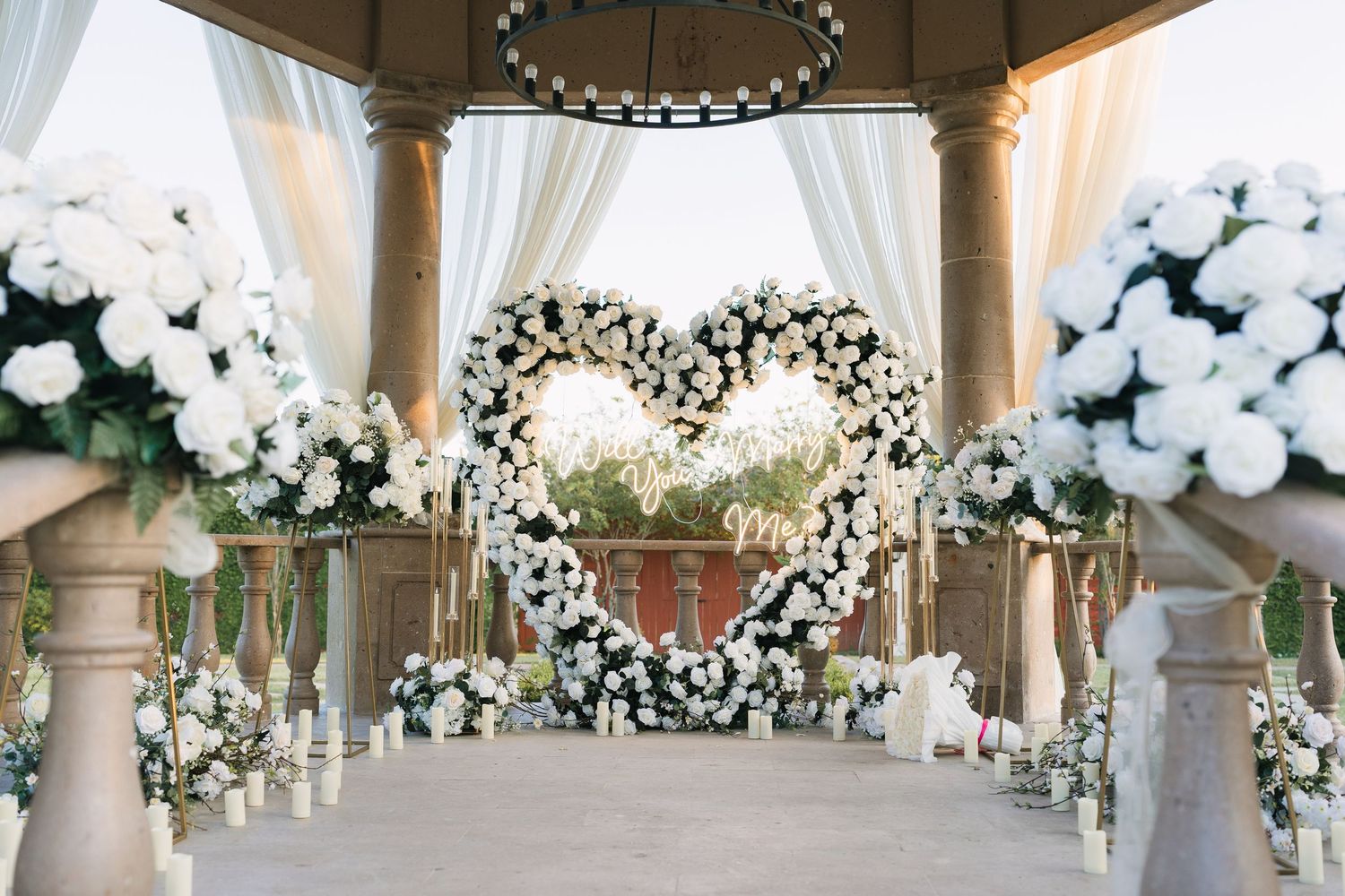 Elegant wedding ceremony with heart-shaped floral arch, white roses, draped fabric, and candles in an outdoor pavilion.