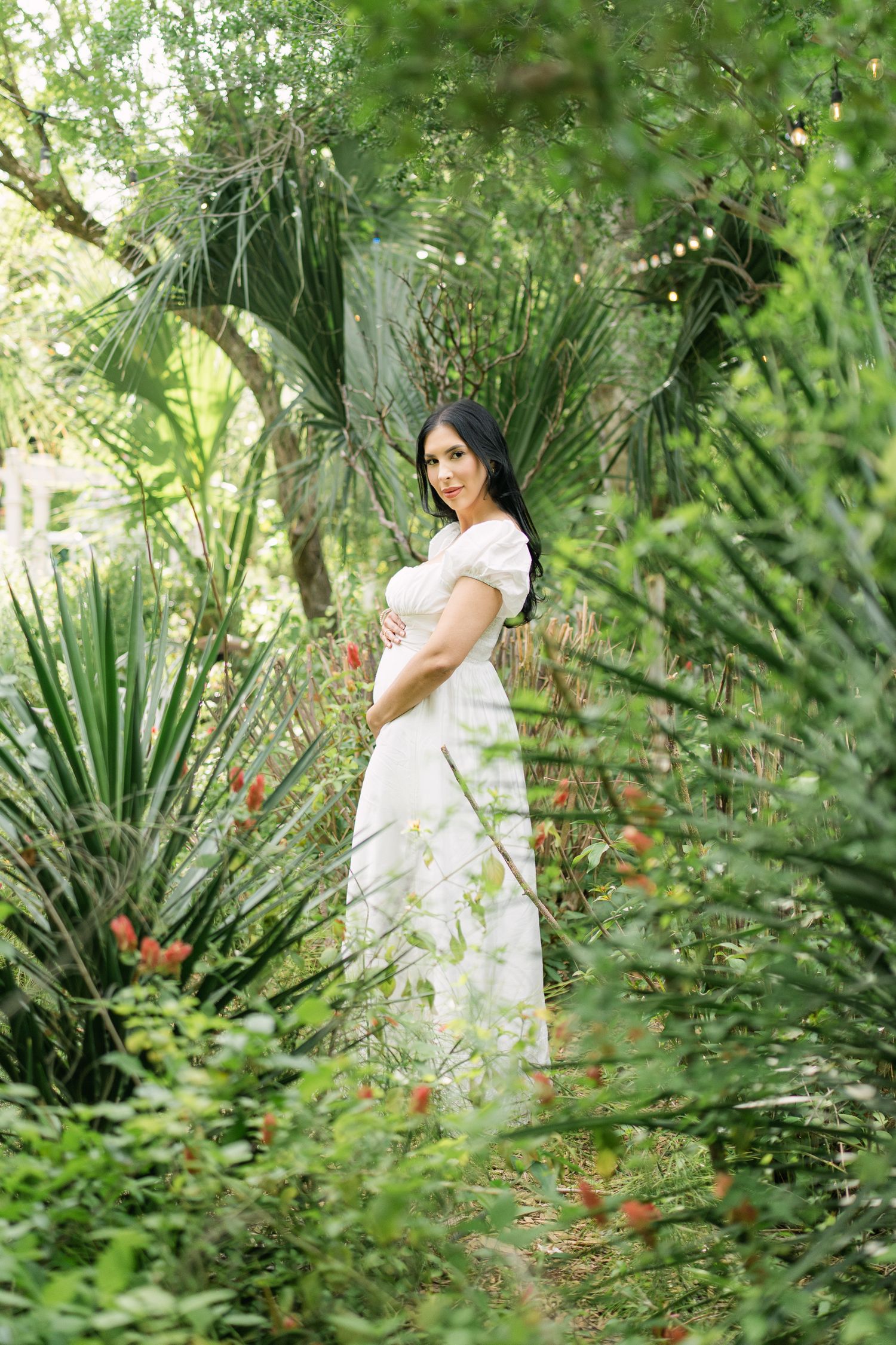 Pregnant woman in white dress poses elegantly among lush tropical garden.