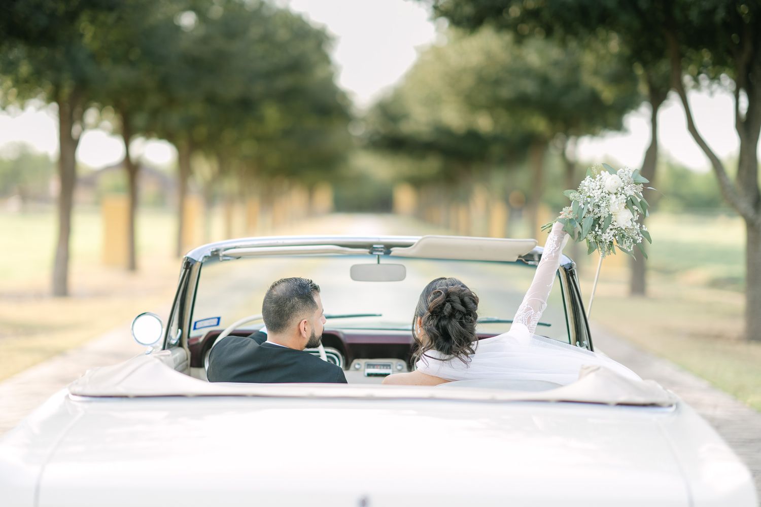 Couple driving away in a vintage convertible on a tree-lined road.