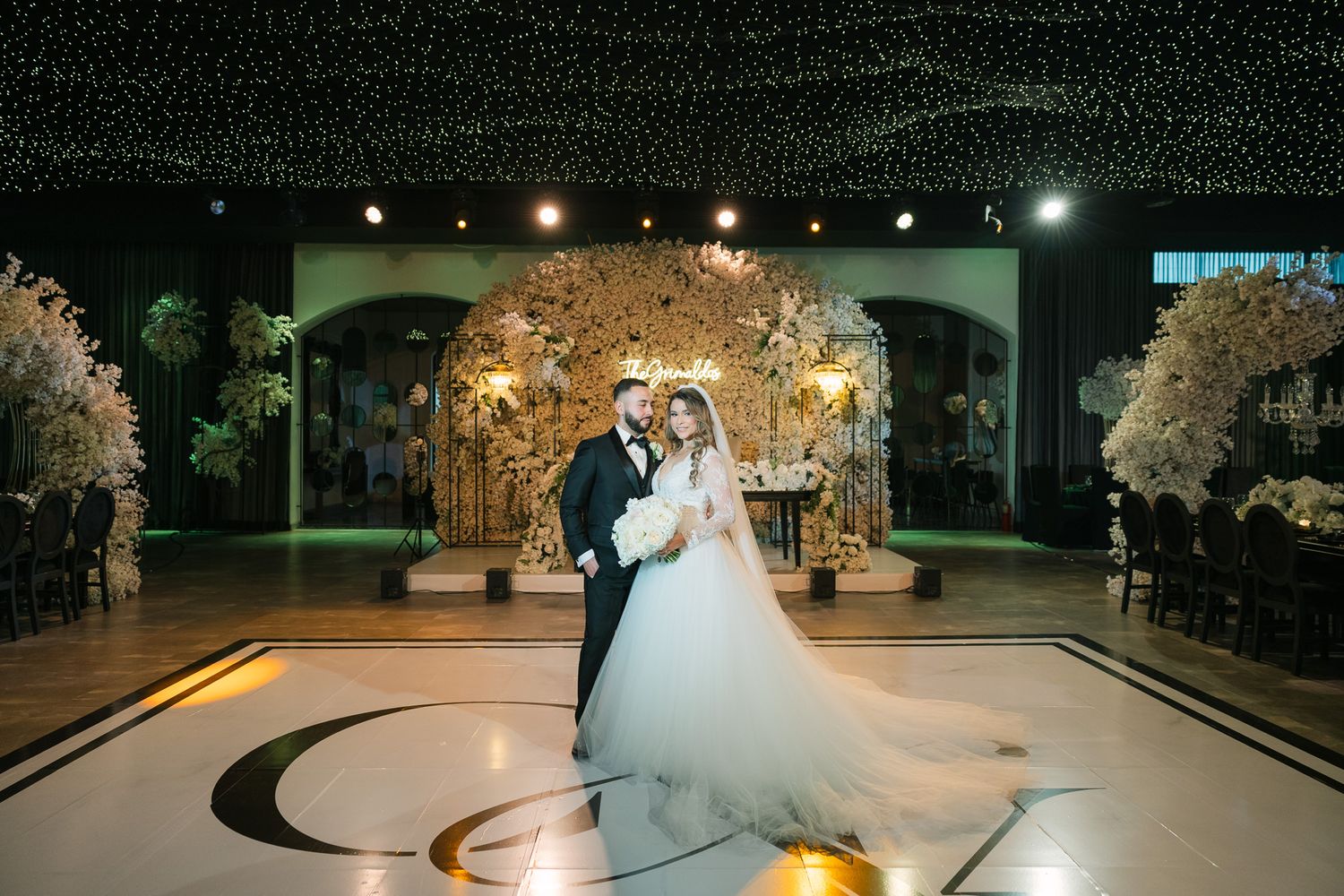 Bride and groom share romantic first dance in elegant ballroom with floral arch backdrop and twinkling ceiling lights.
