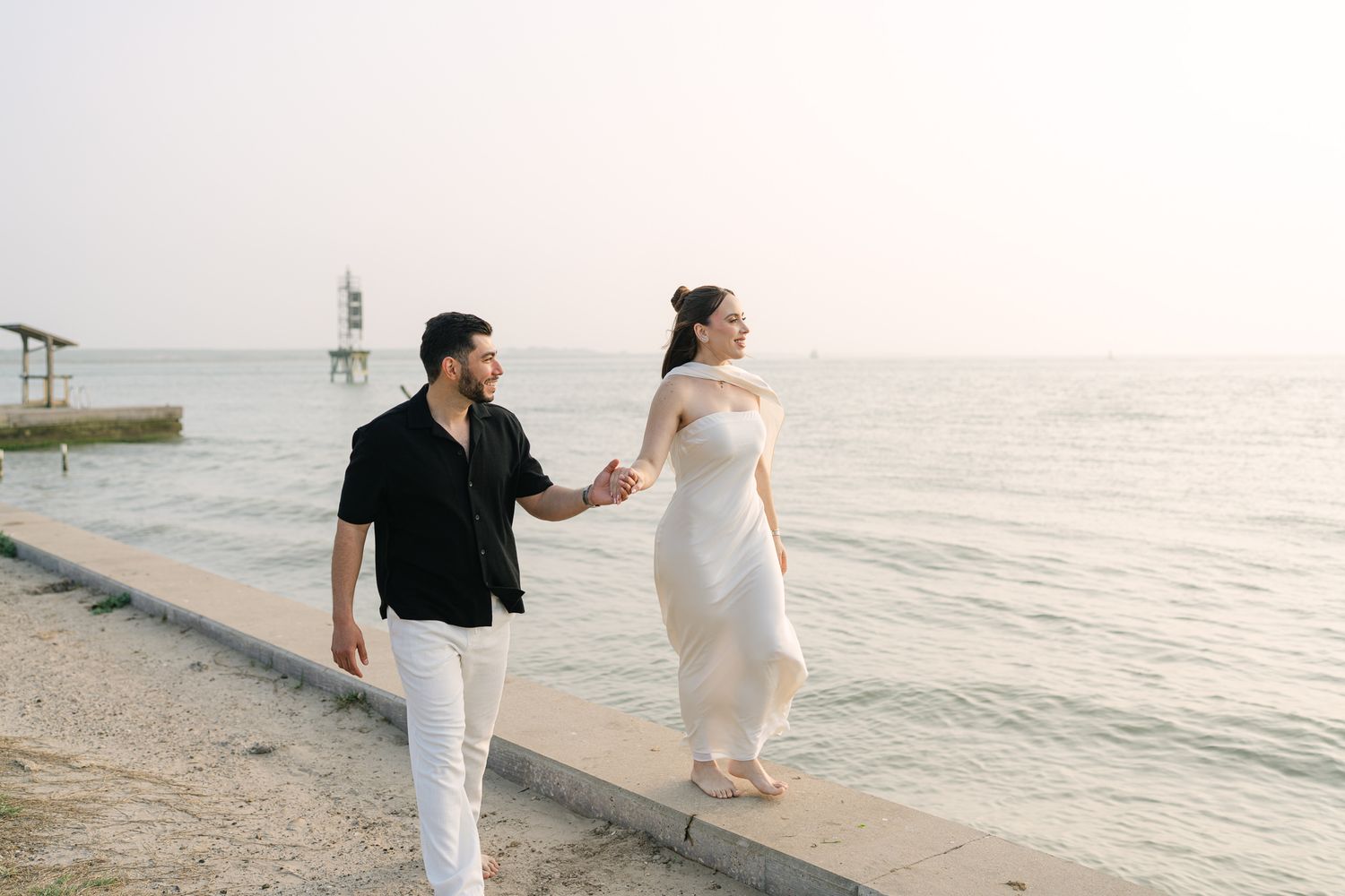 A couple walks hand in hand along a peaceful waterfront at sunset.