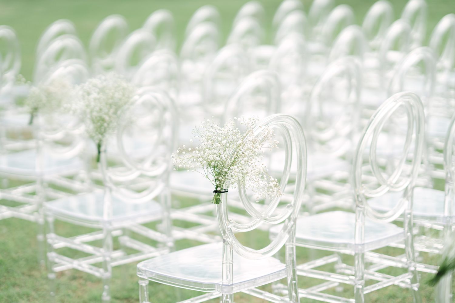 Rows of empty ghost chairs set up for outdoor wedding ceremony with delicate lighting.