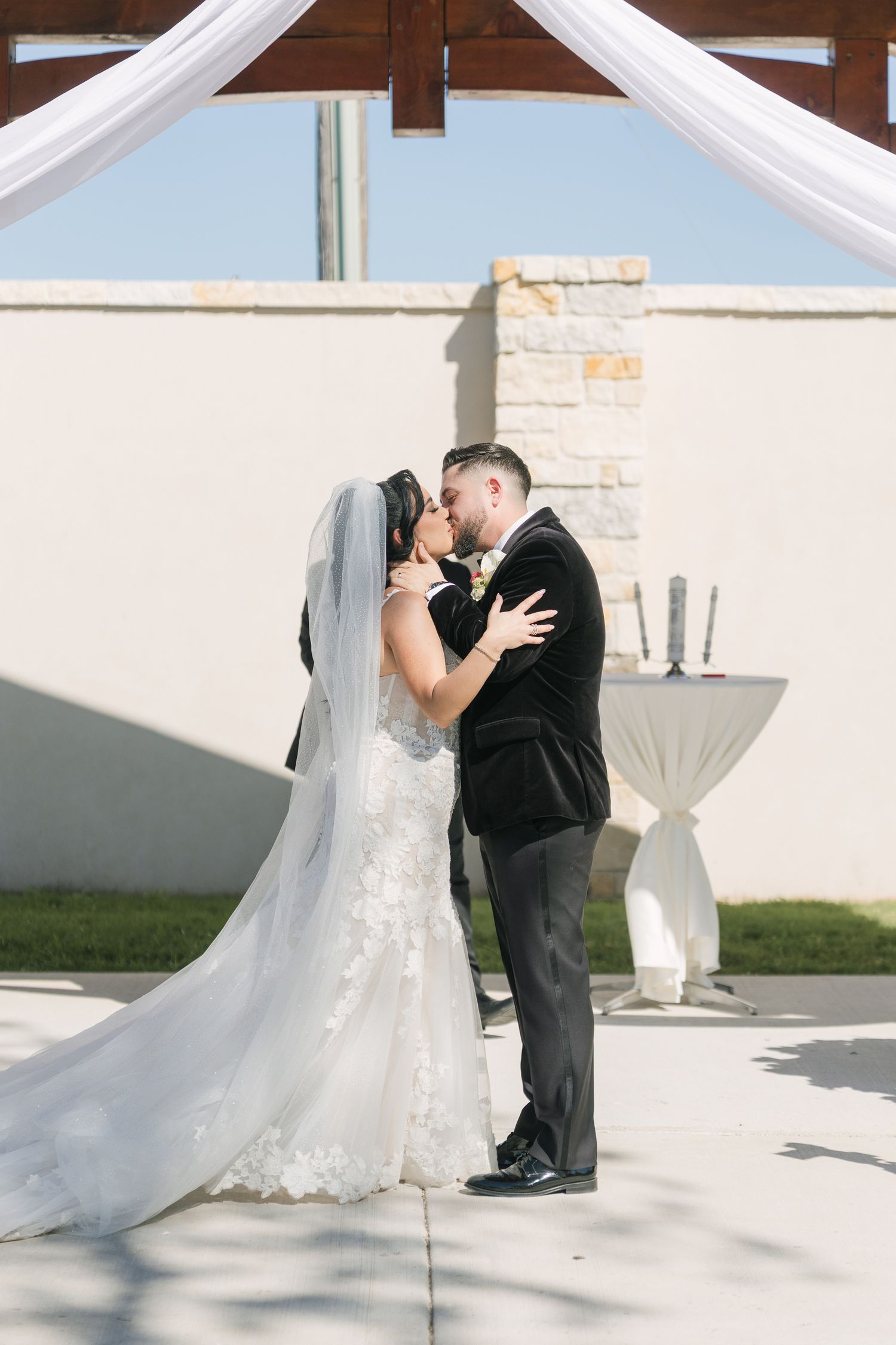 Bride and groom share their first kiss at an outdoor wedding ceremony beneath a white draped arch.