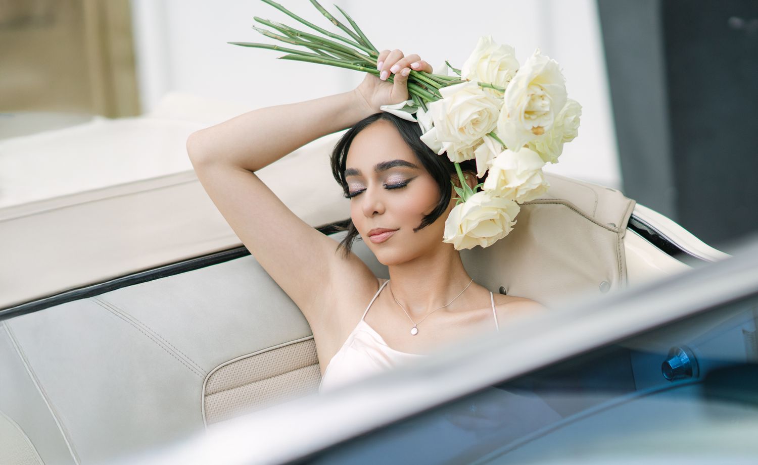 Elegant figure in a blush pink gown reclines in a vintage car with white roses adorning their hair.