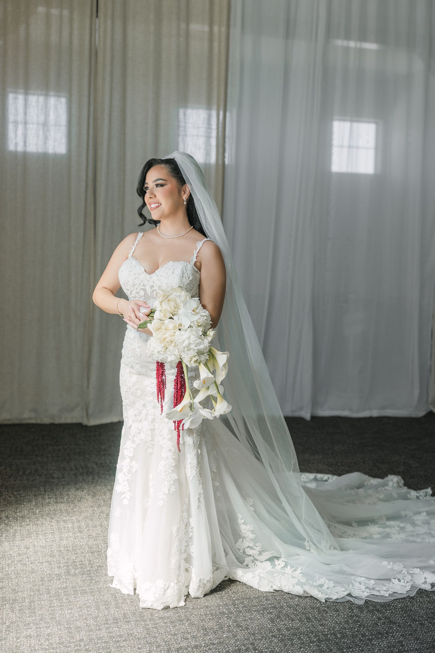 Bride in lace mermaid wedding gown with veil holds white floral bouquet with red ribbon accents, standing by sheer curtains.