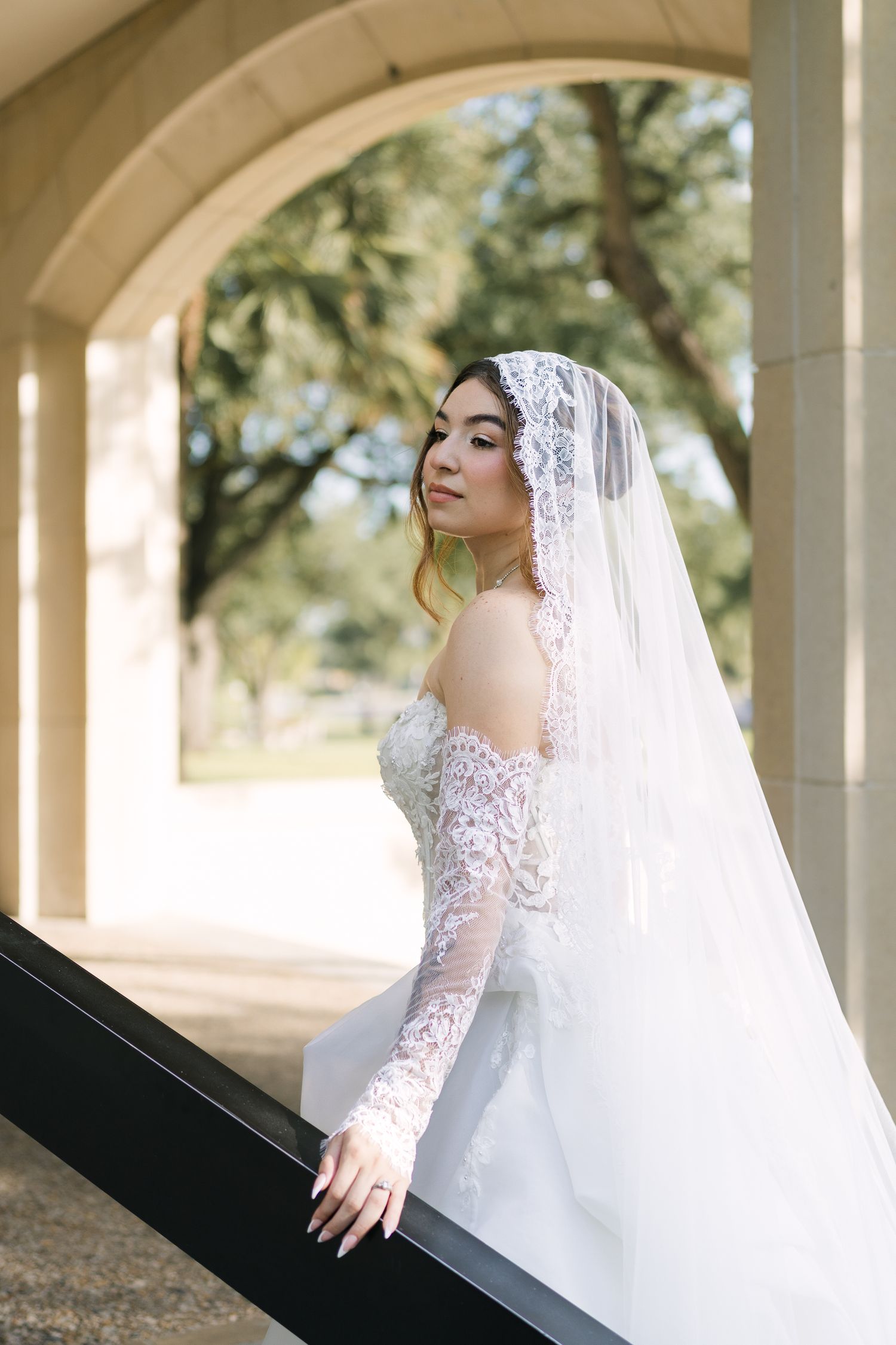 Bride in lace off-shoulder gown and mantilla veil poses gracefully under stone archway surrounded by lush greenery.