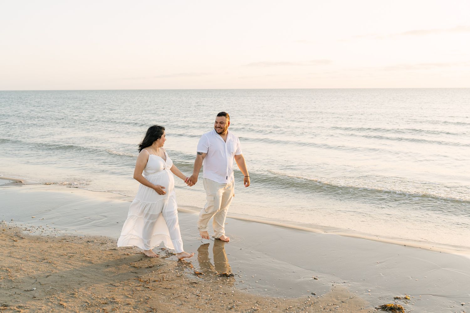 A couple strolling along a beach at sunset in casual white outfits.