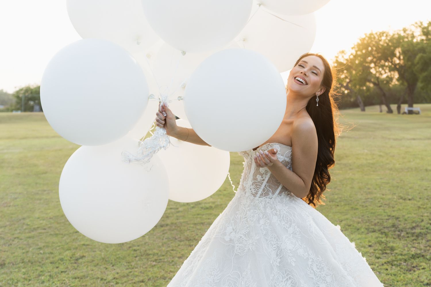 White balloons float against a golden sunset sky during a wedding photoshoot on a grassy hill.