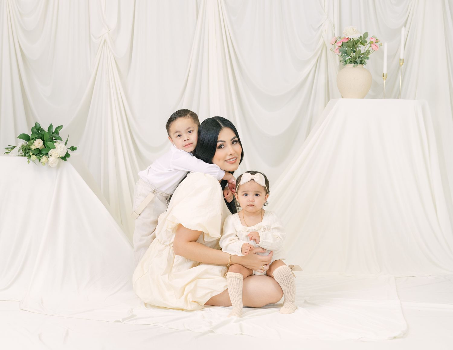 Mother in white dress poses with two children against draped white backdrop.
