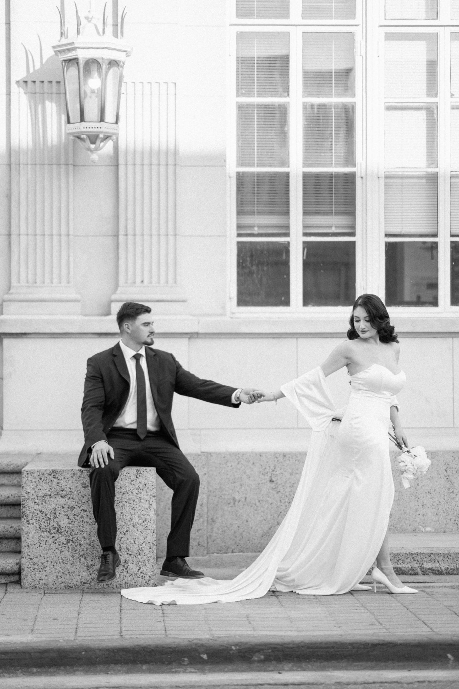 A couple in wedding attire poses playfully while holding hands against a white building exterior.