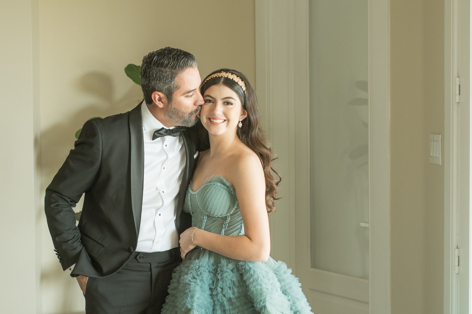 A couple shares intimate moments in formal attire against a light-colored interior setting.