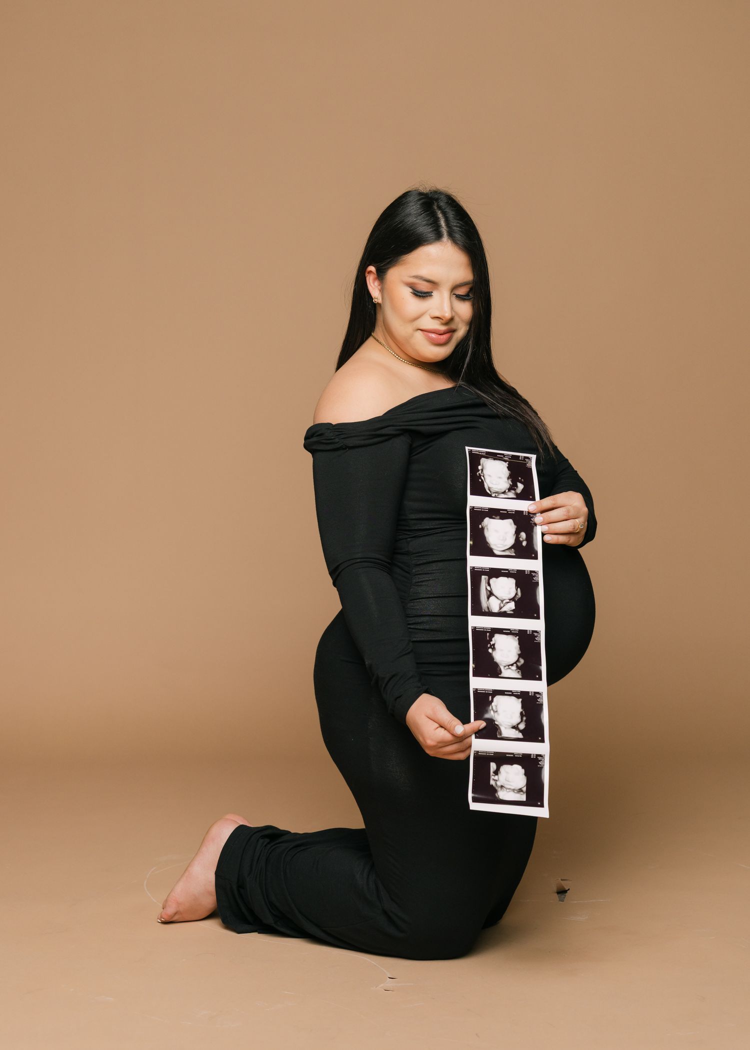 Maternity photoshoot featuring person in black holding ultrasound photos against brown backdrop.