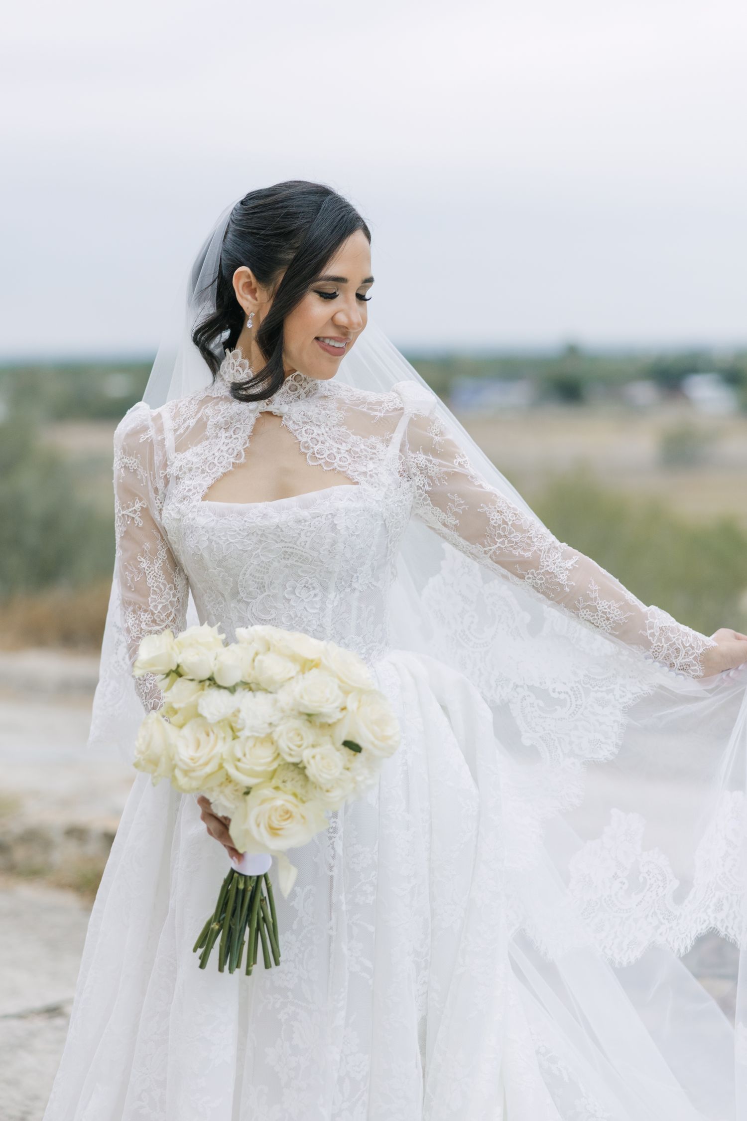 Smiling bride in white lace gown holding bouquet of white roses outdoors on her wedding day.