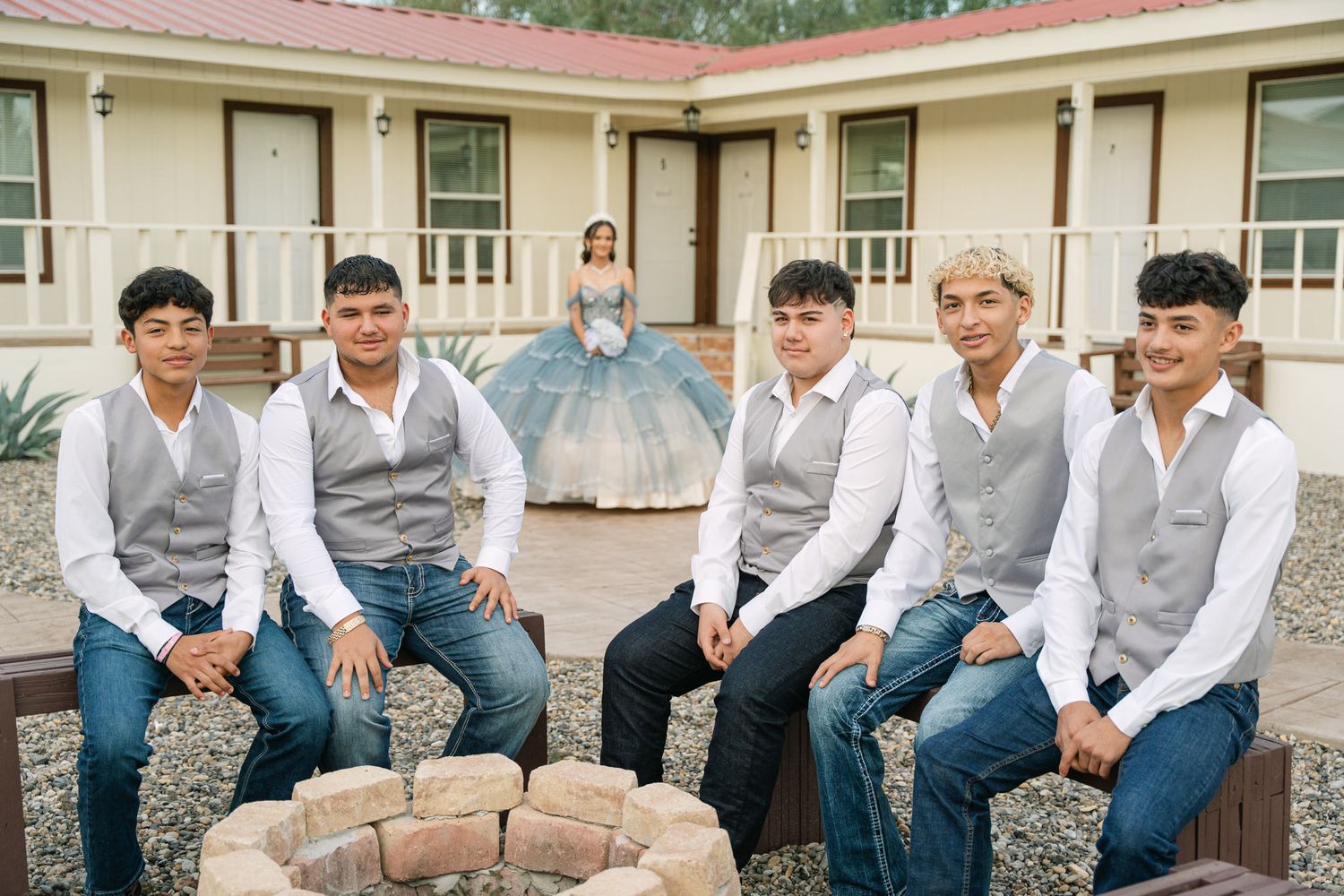 A quinceañera celebrant in a blue dress poses with groomsmen in matching grey vests outdoors.