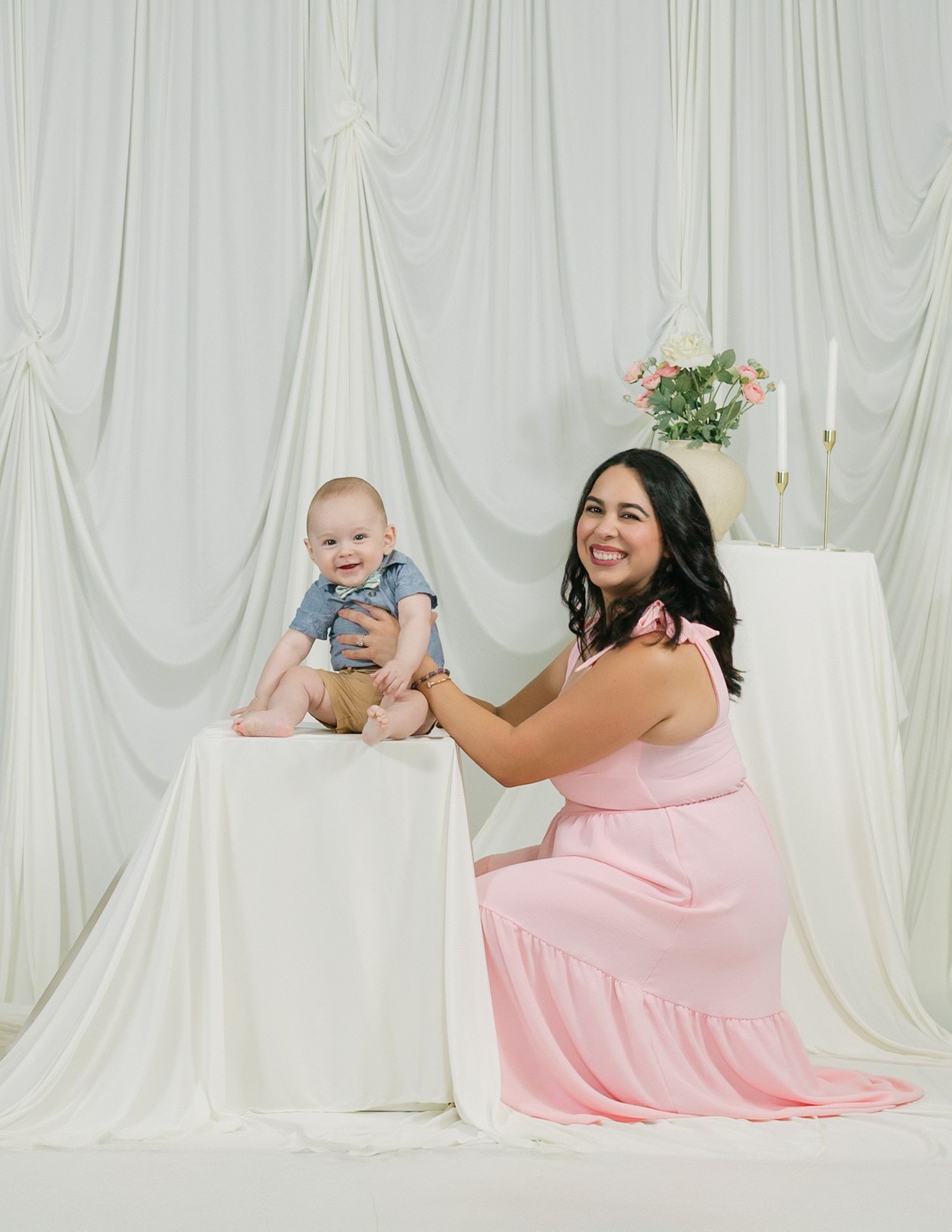 Maternity photo with woman in pink dress posing with baby against white backdrop.