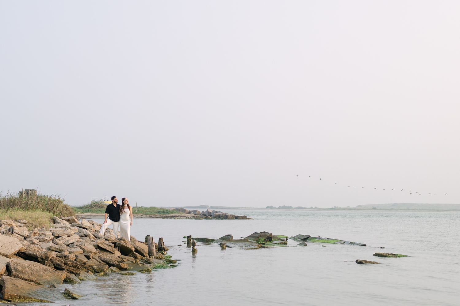 Two silhouettes stand together on rocky coastal shore during golden hour.