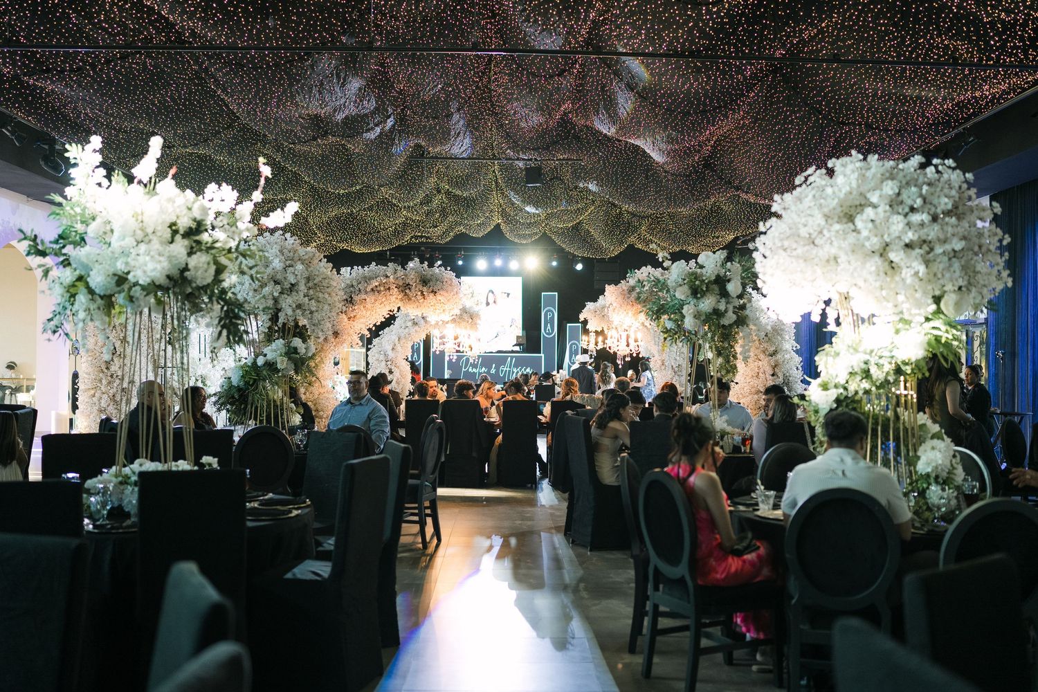 Elegant wedding reception hall with tall white floral arrangements, draped ceiling lights, and guests seated at black-clothed tables.