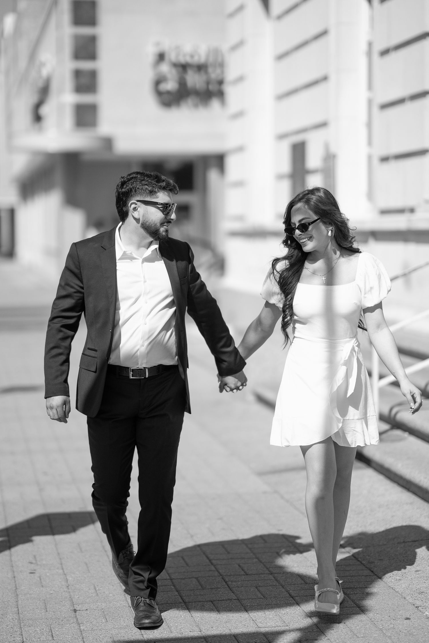 A couple in formal attire walks hand in hand along a sunny urban sidewalk.