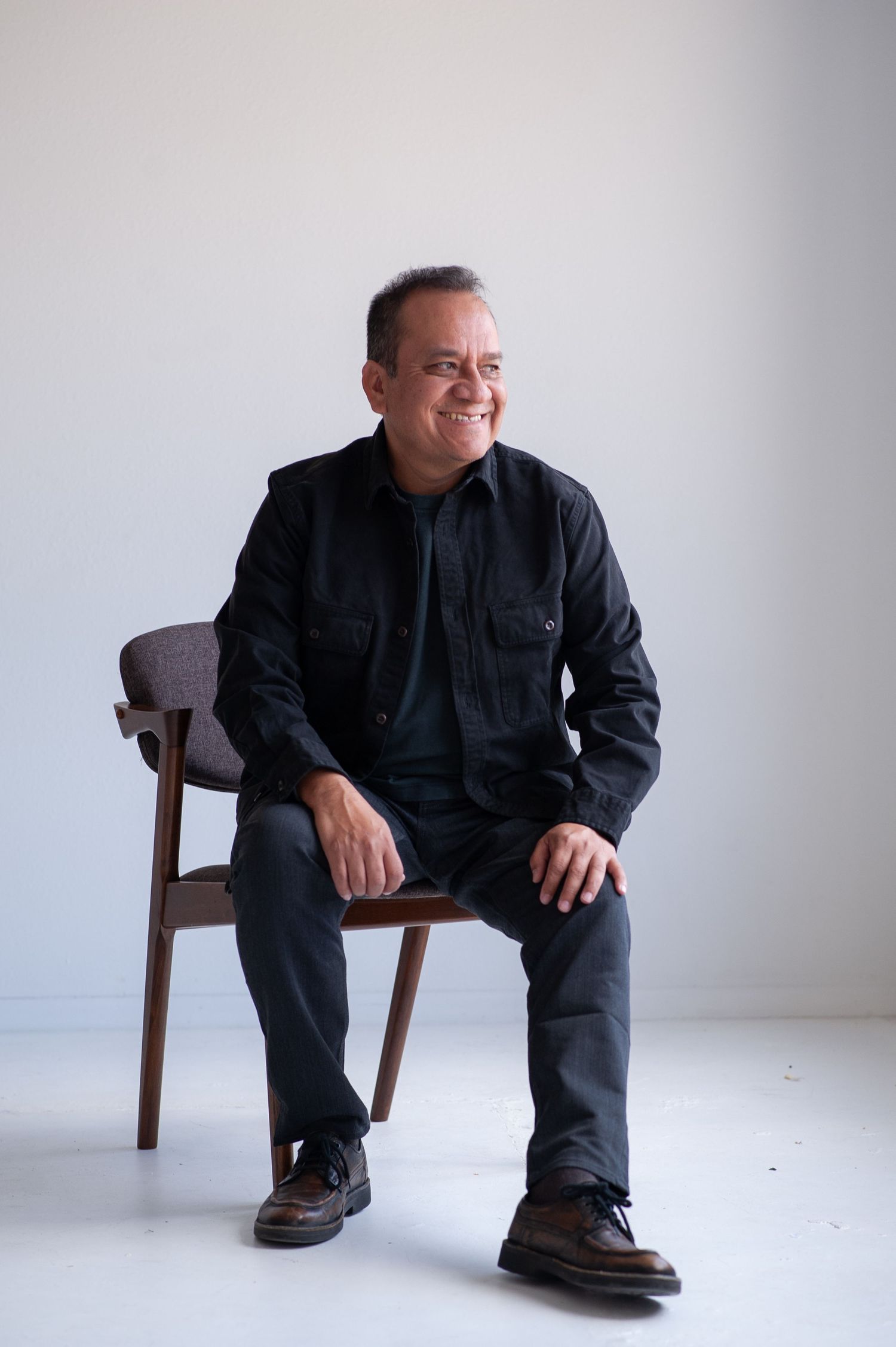 Middle-aged man in black outfit sitting relaxed on wooden chair, smiling against white background.