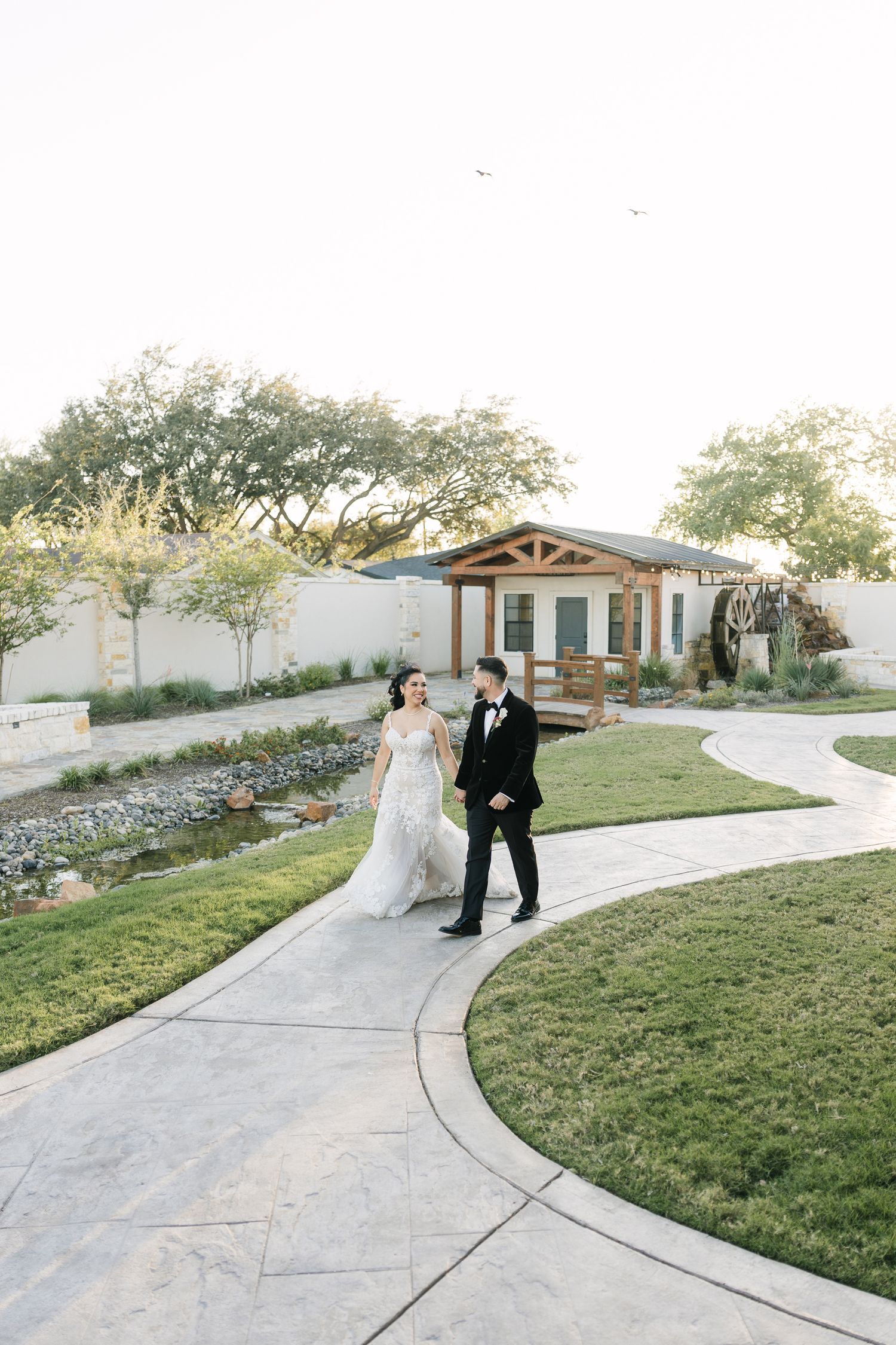 Bride and groom walk hand in hand along a curved garden path at an elegant outdoor wedding venue.