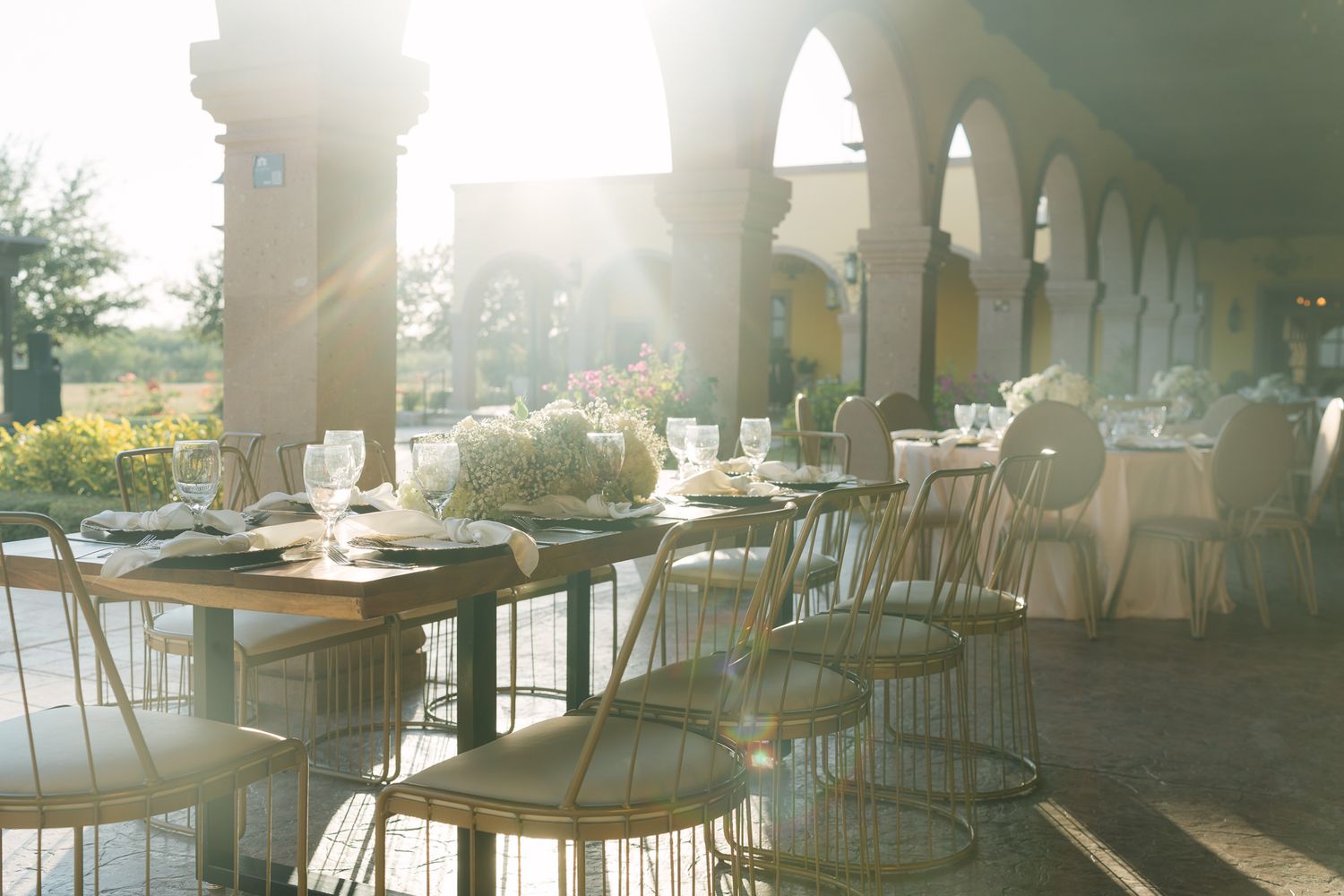 Sunlit outdoor wedding reception with elegant table settings, white floral centerpieces, and arched columns.