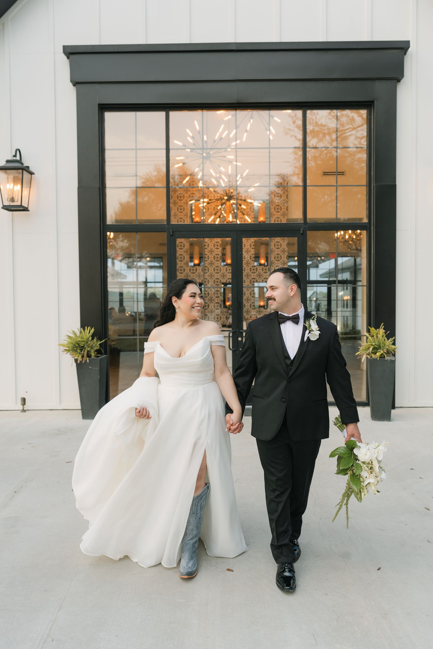 Bride and groom walk hand-in-hand outside a modern white venue, she in an off-shoulder gown, he in a black tuxedo.